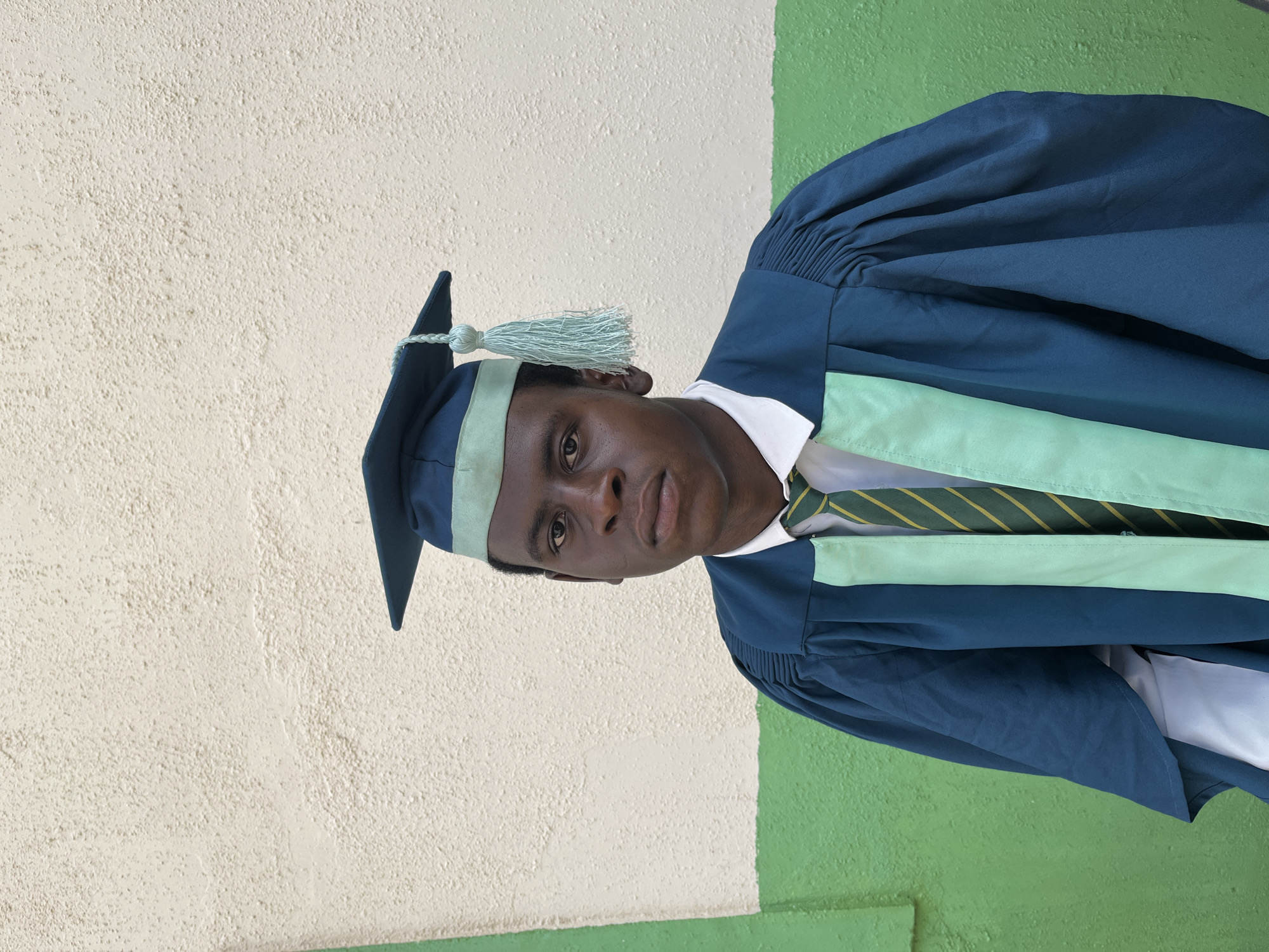 A portrait shows a young man in a graduation cap and gown. The cap is dark blue with a mint green band and tassel. The gown is dark blue with mint green lapels. He is wearing a white shirt and a tie with green and yellow stripes.
He is standing in front of a wall that is painted white on the top portion and green on the bottom portion. The exact location in the city of Jos, Nigeria, cannot be determined from the visual information provided.