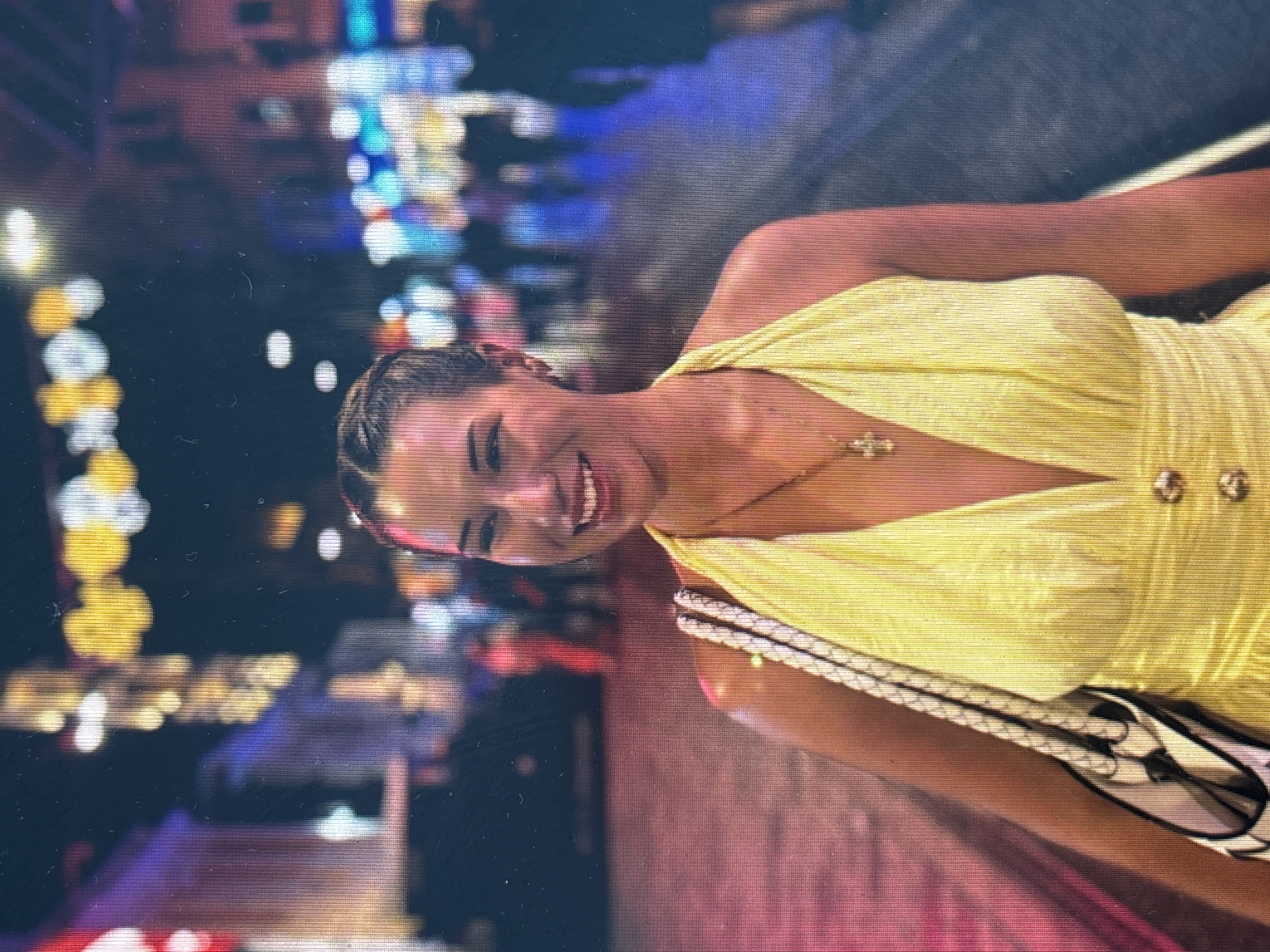 A young woman smiles at the camera, standing in an unidentified location that appears to be a city street at night. She wears a yellow halter dress and a necklace with a cross pendant, and carries a white and black bag over her shoulder. The background features blurred city lights and indistinct structures. No information suggests this is located in Kâğıthane, Turkey.