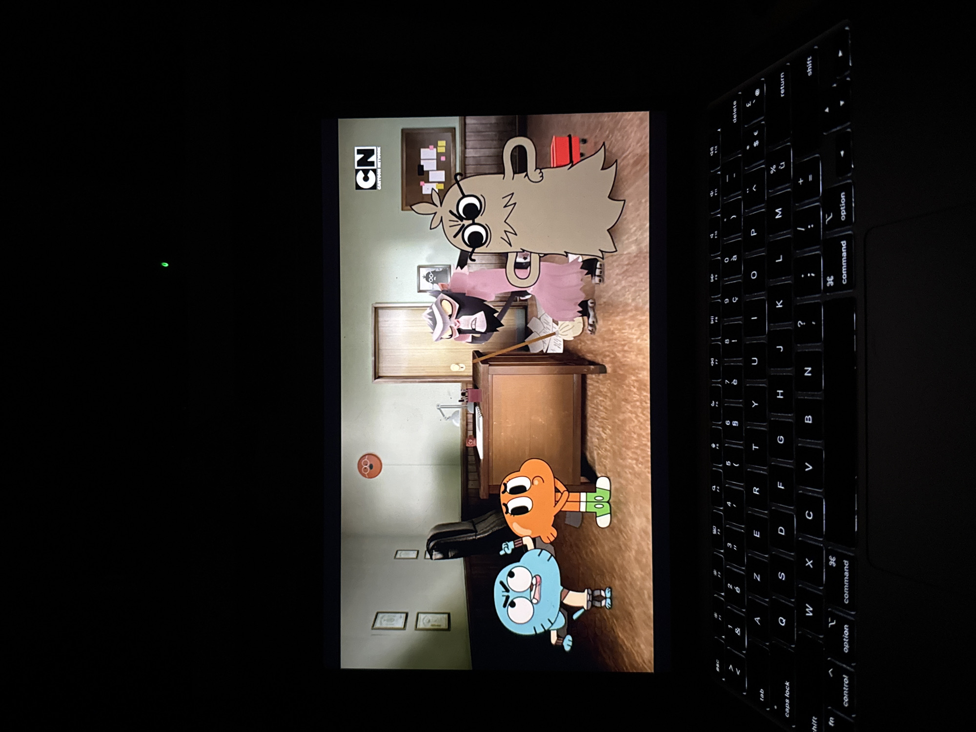 The image shows a laptop screen displaying an episode of "The Amazing World of Gumball." The scene appears to be set inside Principal Brown's office, with Gumball and Darwin standing near his desk. Principal Brown and Ms. Simian are also in the scene. A keyboard is visible to the right of the screen. The laptop is likely located indoors, with the room dimly lit. No location information for Champigny-sur-Marne, France is available from the image.