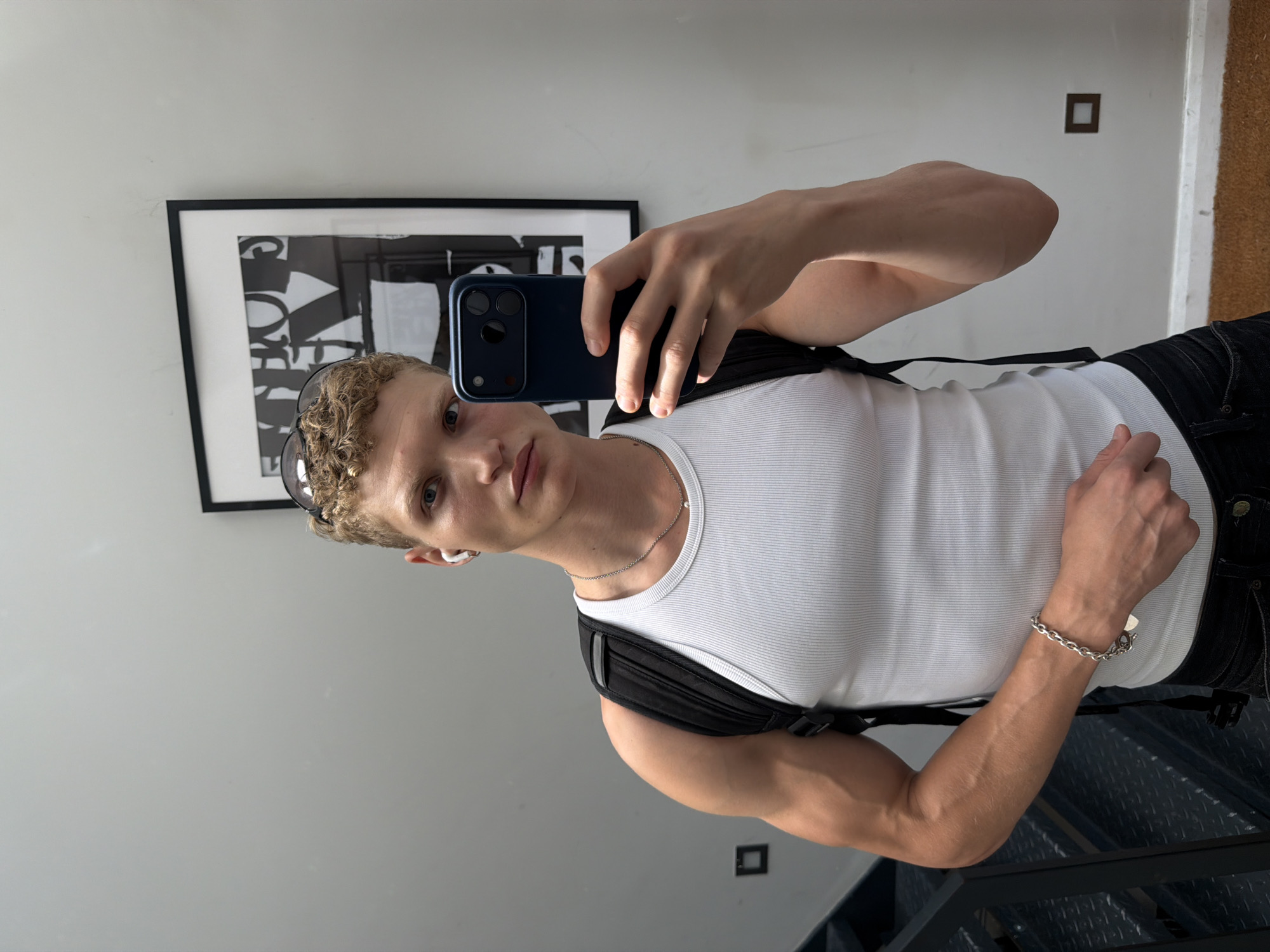 A man with curly blonde hair and blue eyes is taking a selfie in what appears to be an urban setting. He is wearing a white ribbed tank top, a black backpack, dark jeans, a silver chain necklace, a silver bracelet, and white wireless earbuds. Sunglasses are perched on his head. He is holding a dark blue smartphone in his right hand to take the photo. Behind him, a large framed black and white artwork with bold lettering hangs on a light grey wall. To his left, a metal staircase with diamond plate treads descends. The overall environment suggests a modern, possibly loft-style interior with a minimalist aesthetic. There is no specific information within the image to determine the city as London, United Kingdom.