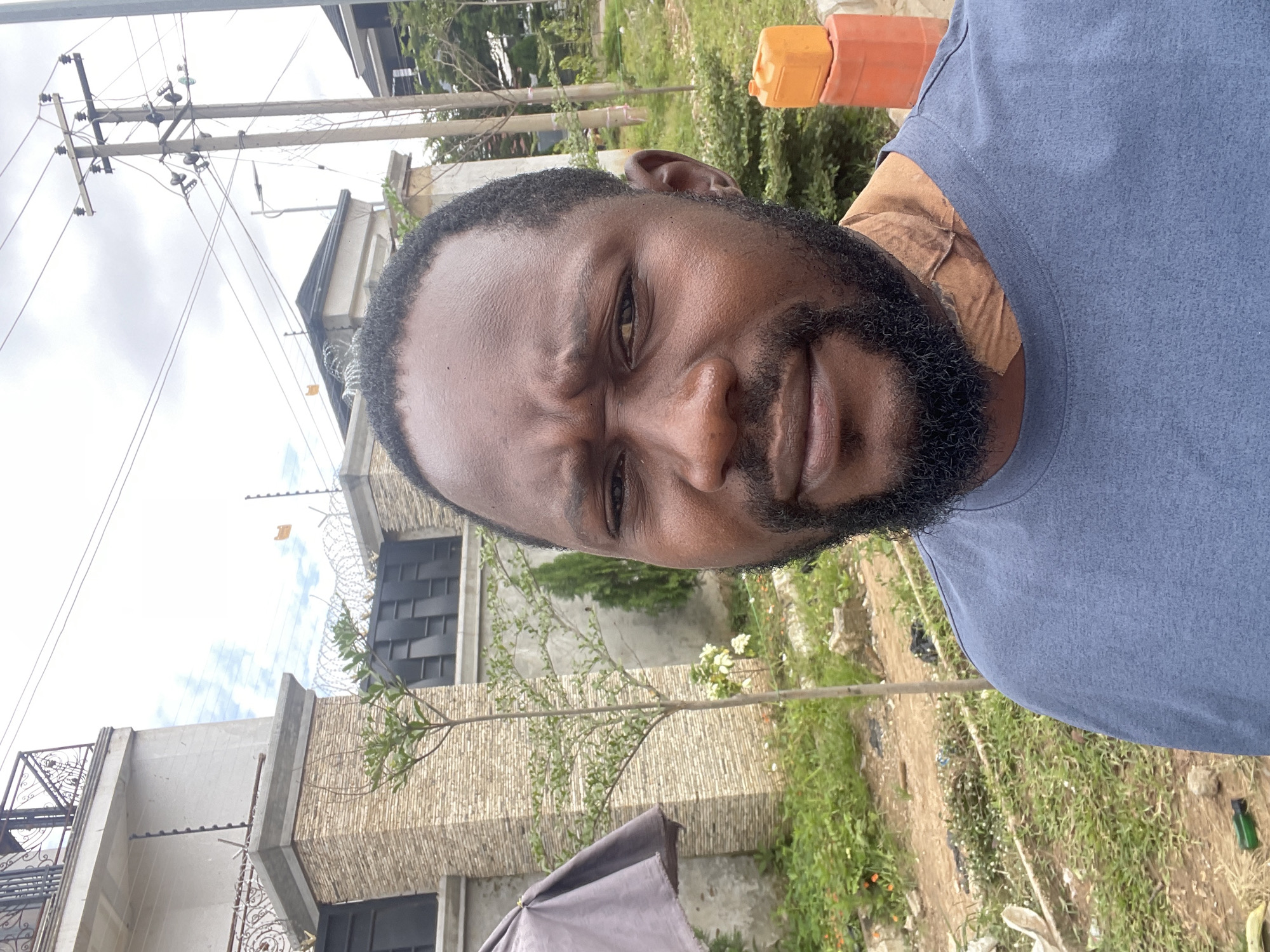A man with dark skin, a beard, and a blue t-shirt is captured in a close-up, eye-level selfie. He has a stern expression and his right eye is slightly squinted. A piece of light brown material is wrapped around his neck. He is standing outdoors in what appears to be a construction or developing area. In the background, there is a building with stone accents and a metal fence with barbed wire. Further back, there are power lines and utility poles, characteristic of a city environment. To the right, there are stacked orange plastic containers. The ground is covered with dirt, grass, and some debris. This scene is located in Guzape, Nigeria.