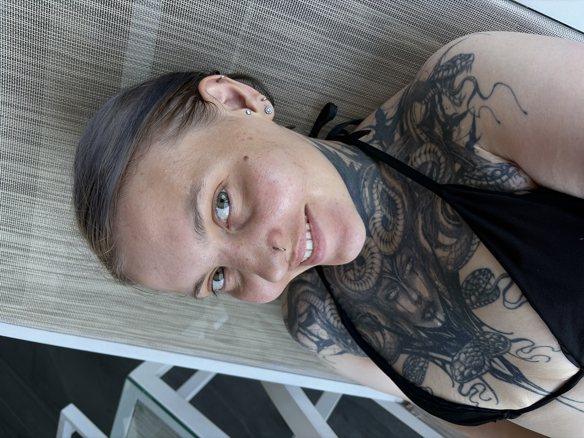 A woman reclines on a light-colored mesh lounge chair, captured from a high-angle perspective, looking up towards the camera with a relaxed smile. Her head is positioned to the left, and her body extends towards the right of the frame. She has dark hair, slicked back from her face, and light-colored eyes.
Her face features a nostril ring and multiple ear piercings, including a helix ring, a stud, and a floral-patterned stud. She has clear skin with a few freckles.
A prominent feature is the extensive blackwork tattooing covering her chest, left shoulder, and neck. The tattoos depict intricate designs including snakes, swirling patterns, and what appears to be a stylized face or bust, possibly Medusa-like, in the center of her chest. She is wearing a black bikini top that partially covers the tattoos on her chest.
The background shows the textured mesh of the lounge chair, and in the lower right, part of a white table or chair frame is visible against a darker, possibly tiled or wooden, floor. The lighting is bright and natural, suggesting it is daytime and likely outdoors, contributing to a relaxed atmosphere. The overall scene implies a leisurely setting, possibly at a resort or by a pool in Đà Nẵng, Vietnam. There is no visible text in the image.