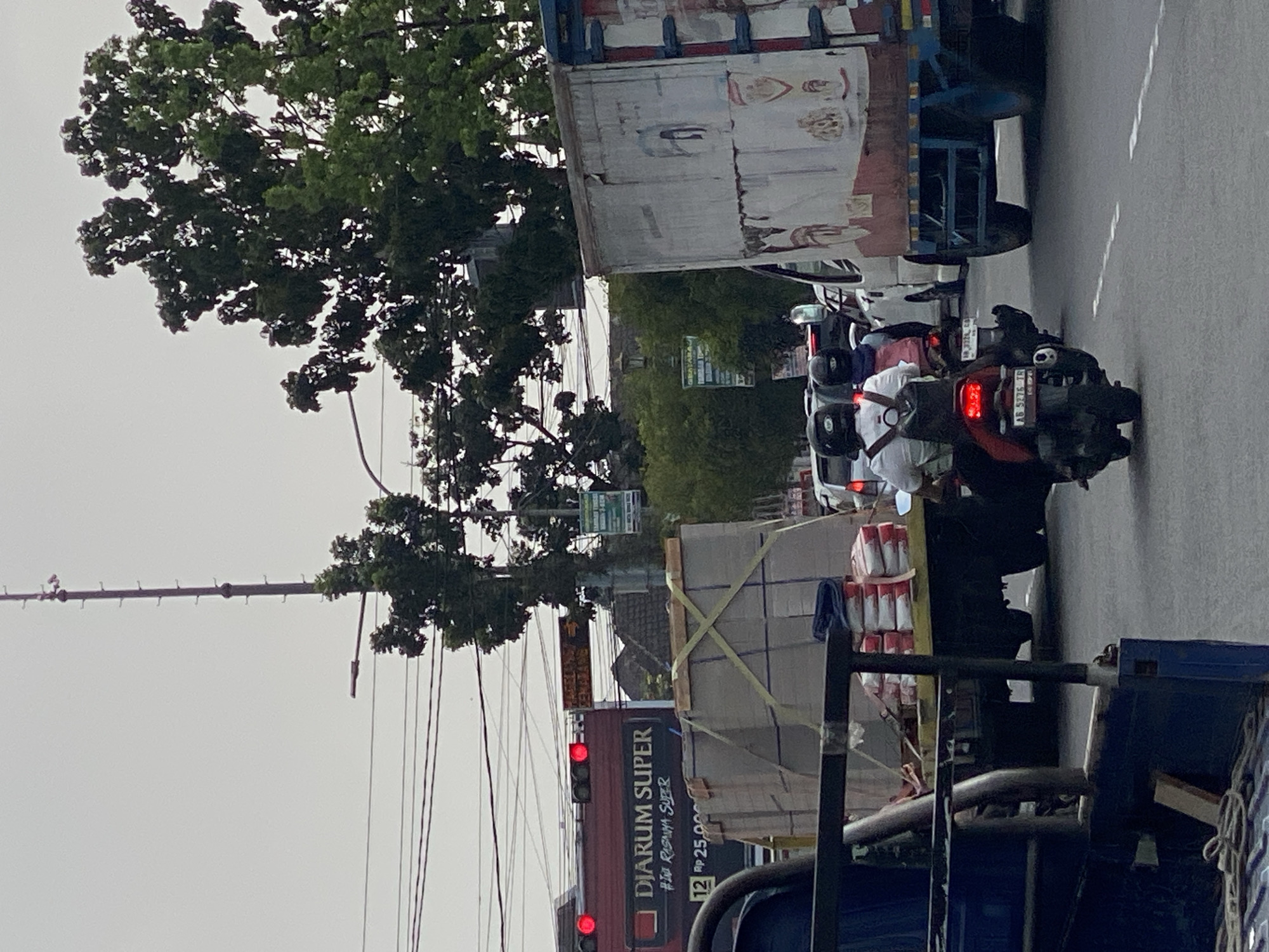 The image depicts a street scene in Tamantirto, Indonesia, with heavy traffic under an overcast sky. A large red truck with "DJARUM SUPER" branding and a price of "12 Rp 25.000" is visible on the left, its brake lights illuminated. Beside it, a flatbed truck carries large, wrapped rectangular objects and stacks of bagged materials. In the foreground, two individuals on a motorcycle are navigating through traffic. The rider wears a white shirt and a black backpack, while the passenger has a pink backpack. The motorcycle's license plate is partially visible. Further ahead, a white truck with faded advertisements on its side is also part of the traffic flow. Trees line the street, and power lines crisscross the sky. The environment suggests a busy urban or semi-urban area.