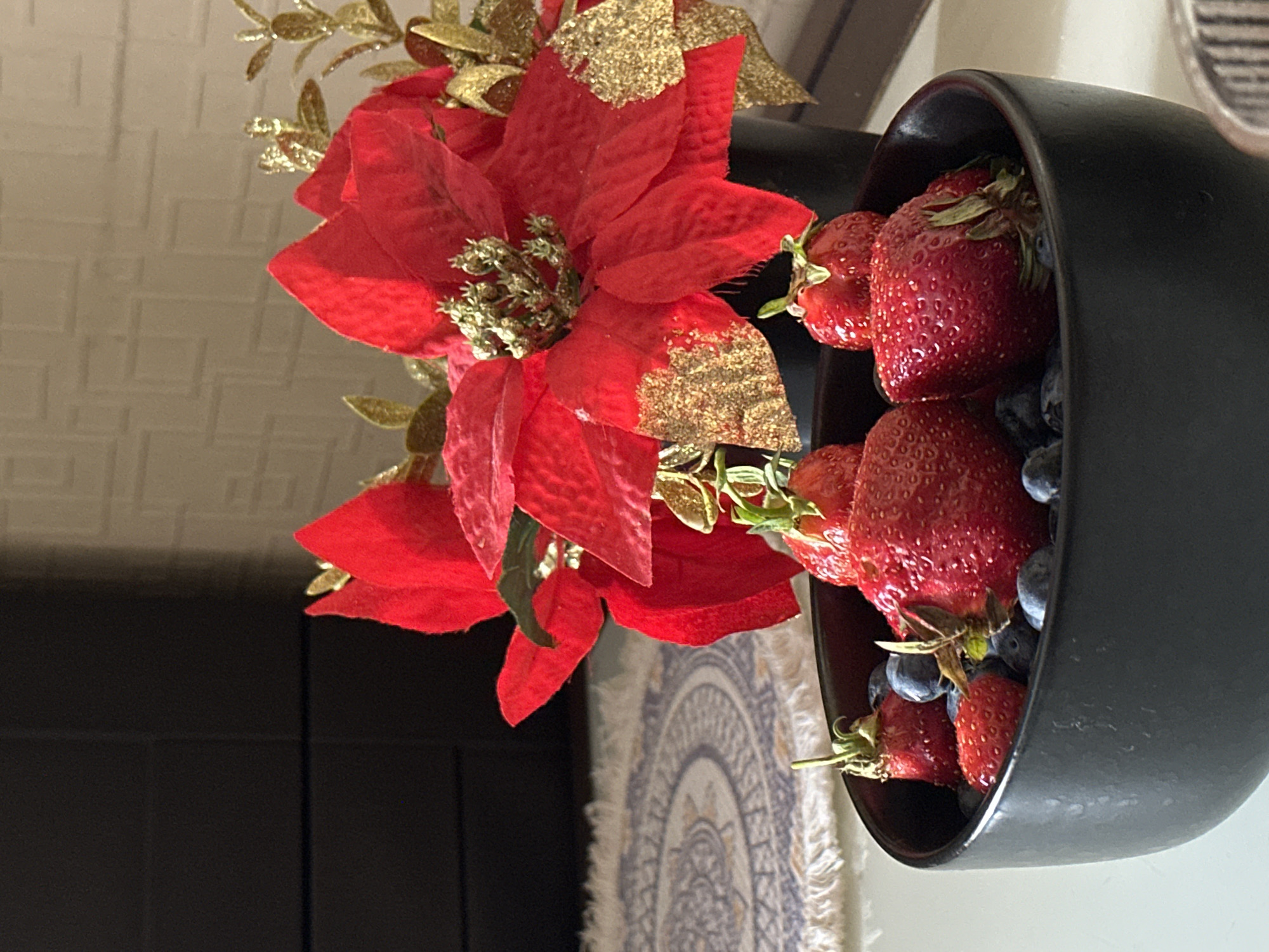 A close-up, eye-level shot captures a dark black bowl filled with fresh strawberries and blueberries. The fruit is glistening, suggesting it might be slightly wet or very ripe. Several large, red strawberries are prominent, some with green stems still attached. The blueberries are scattered amongst the strawberries, appearing smaller and a deep blue color.
Behind the bowl, a bouquet of artificial poinsettias stands tall. The poinsettias are a vibrant red, with some petals adorned with glittery gold accents. They are arranged in a black pot, partially visible.
The background is a bit out of focus. To the left, a dark, rectangular surface, possibly a piece of furniture or a screen, is visible. To the right and slightly behind the poinsettias, a textured, light-colored wall with a subtle pattern can be seen. A portion of a rug with a circular, intricate design and fringed edges is visible in the lower left corner, resting on a light-colored surface.
The lighting suggests an indoor setting, possibly with natural light coming from the right, casting subtle shadows and highlighting the textures of the fruit and flowers. The overall scene evokes a sense of a holiday or festive occasion, perhaps a breakfast setup. There are no people present in the image, and no specific details about the city of Kyiv, Ukraine are discernible from the visual information provided.