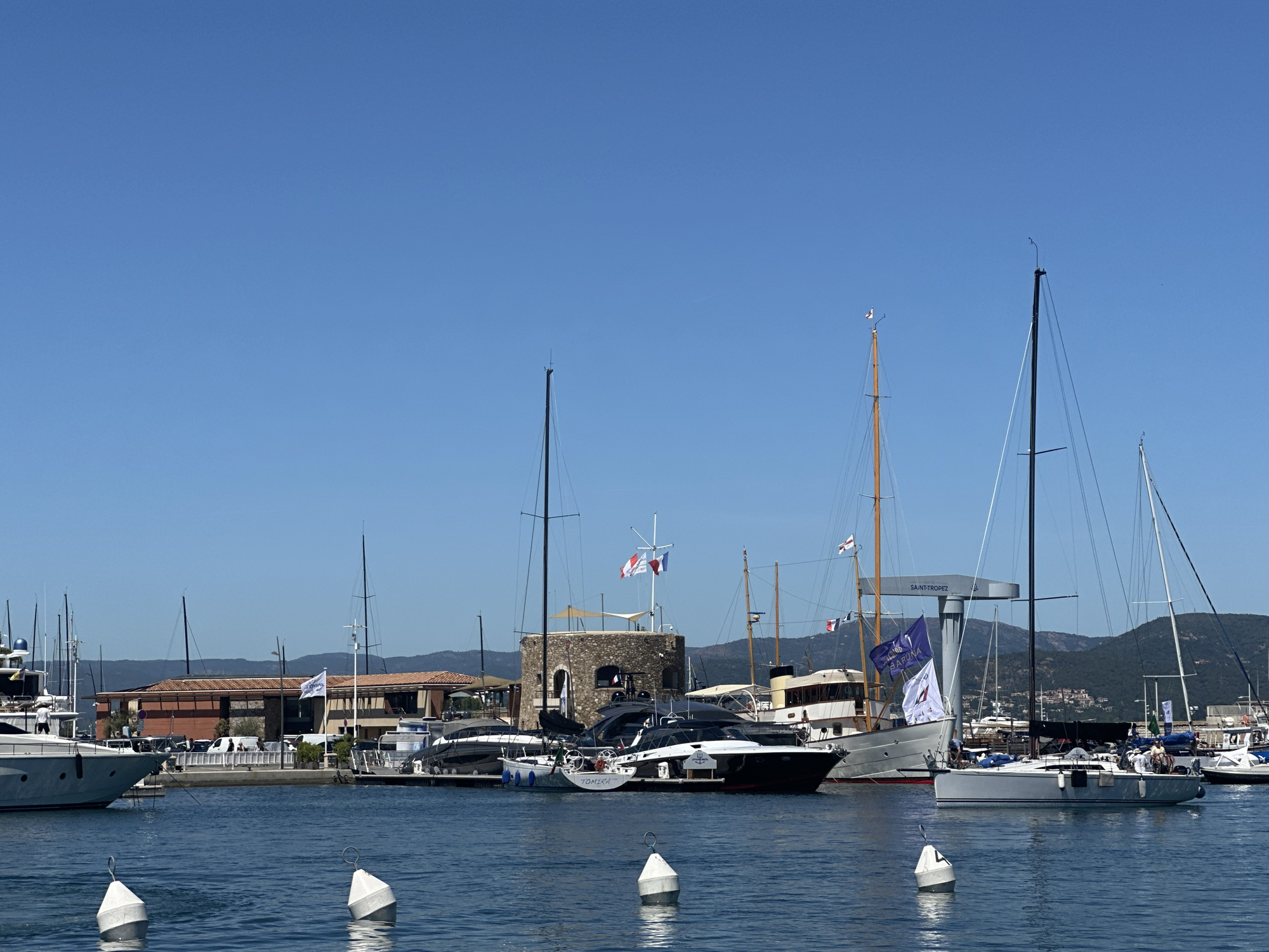 The image shows a harbor in Saint-Tropez, France, with several yachts and sailboats docked in the blue water under a clear sky. In the foreground, white buoys bob on the water. A large building with an orange-tiled roof and a stone tower is visible behind the boats. The boats range in size and design, with some appearing to be luxury yachts. Some of the boats have flags flying from their masts, including the French flag and what appears to be a club or event flag. In the background, green hills rise behind the town. A few people are visible on the docks and on some of the boats, but their actions are not discernible. The overall scene depicts a sunny day in a busy and scenic harbor.