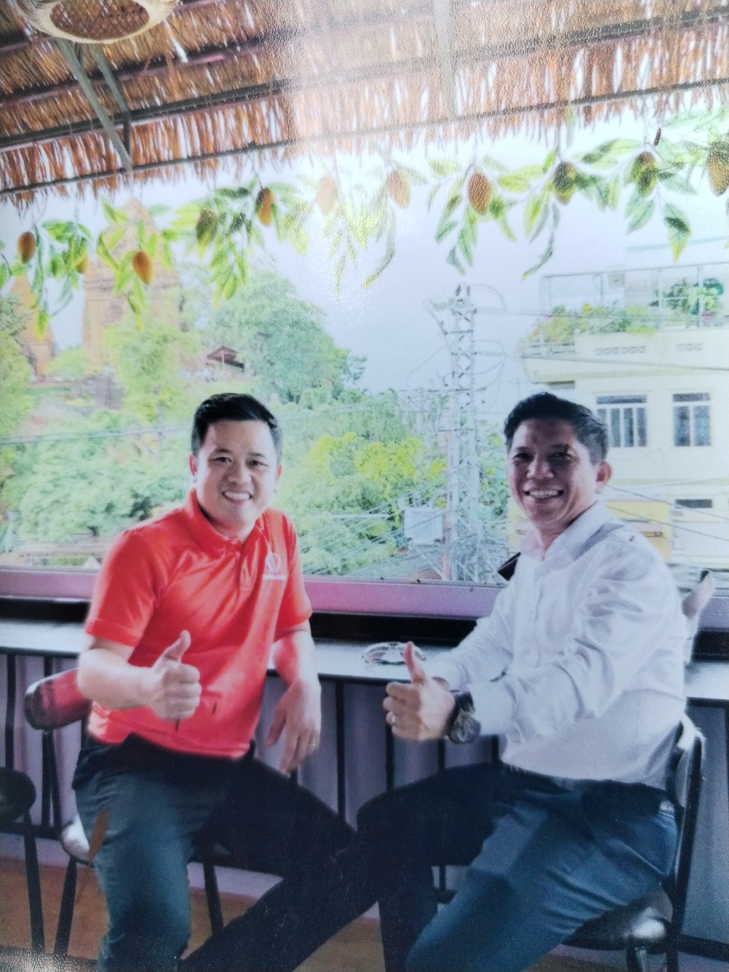 Two men are seated outdoors, giving thumbs-up gestures. The man on the left wears a red polo shirt and dark pants. The man on the right wears a white long-sleeved shirt and dark pants. They are seated in front of a backdrop depicting a cityscape with trees and buildings. Above them is a thatched roof. The location is identified as Bac Nha Trang Ward, Vietnam.