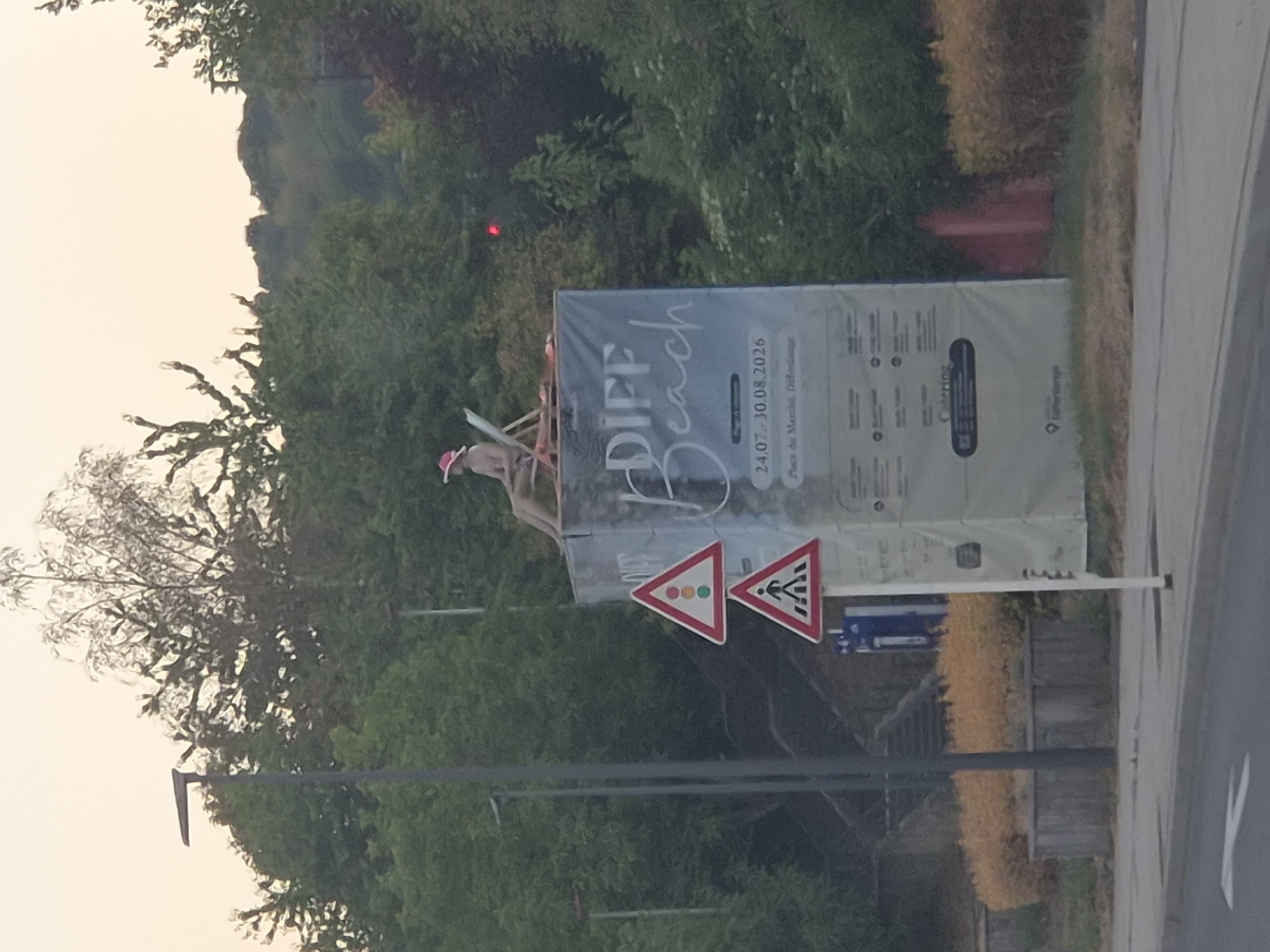 A static, medium-wide outdoor shot shows a large banner for "DIFF Beach" positioned in the city of Differdange, Luxembourg, with green trees in the background and a street in the foreground. The banner is a light color with dark text and images. The text reads "DIFF Beach" in large, stylized lettering. Below this, it displays dates "24.07 - 30.08.2026" and "Place du Marché, Differdange". At the top of the banner, a person is sitting on a beach chair, wearing a pink hat and seemingly sunbathing. Two traffic signs are visible in front of the banner: one triangular sign with a traffic light symbol and another triangular sign depicting a pedestrian crossing. A streetlamp is positioned to the left of the banner. The foreground features a grey paved surface with a white directional arrow, and a concrete planter with dry, orange-colored shrubs. The sky is overcast with a light yellow hue.