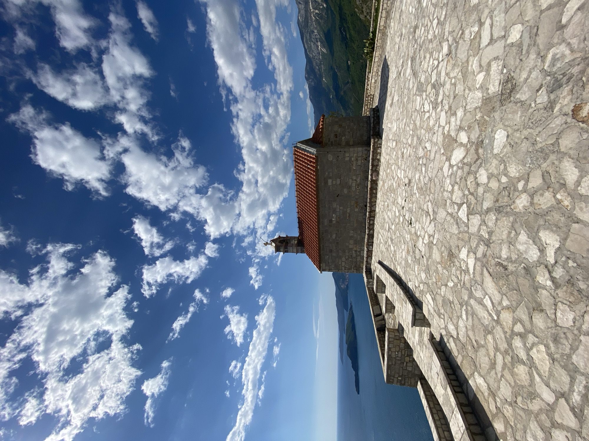 The image captures a picturesque coastal scene, likely in Sveti Stefan, Montenegro, under a bright blue sky dotted with numerous fluffy white clouds. In the foreground and midground, a small, stone building with a red-tiled roof stands prominently. A delicate bell tower with a cross adorns the roof of this structure, which appears to be a chapel or church.
The building is situated on a wide, irregularly paved stone terrace or walkway, made of various shades of light gray and beige stones. To the right of the building, a low, curved stone balustrade or wall provides a boundary for the terrace.
Beyond the architectural elements, a vast expanse of calm, deep blue water stretches towards the horizon. An island or peninsula, green with vegetation, rises from the water, enhancing the scenic beauty. In the far background, majestic green-clad mountains loom, completing the dramatic landscape.
The time of day appears to be sunny daytime, with clear visibility and bright illumination, casting subtle shadows. There are no people visible, and no discernible text is present in the image. The overall impression is one of serene natural beauty and historic charm.