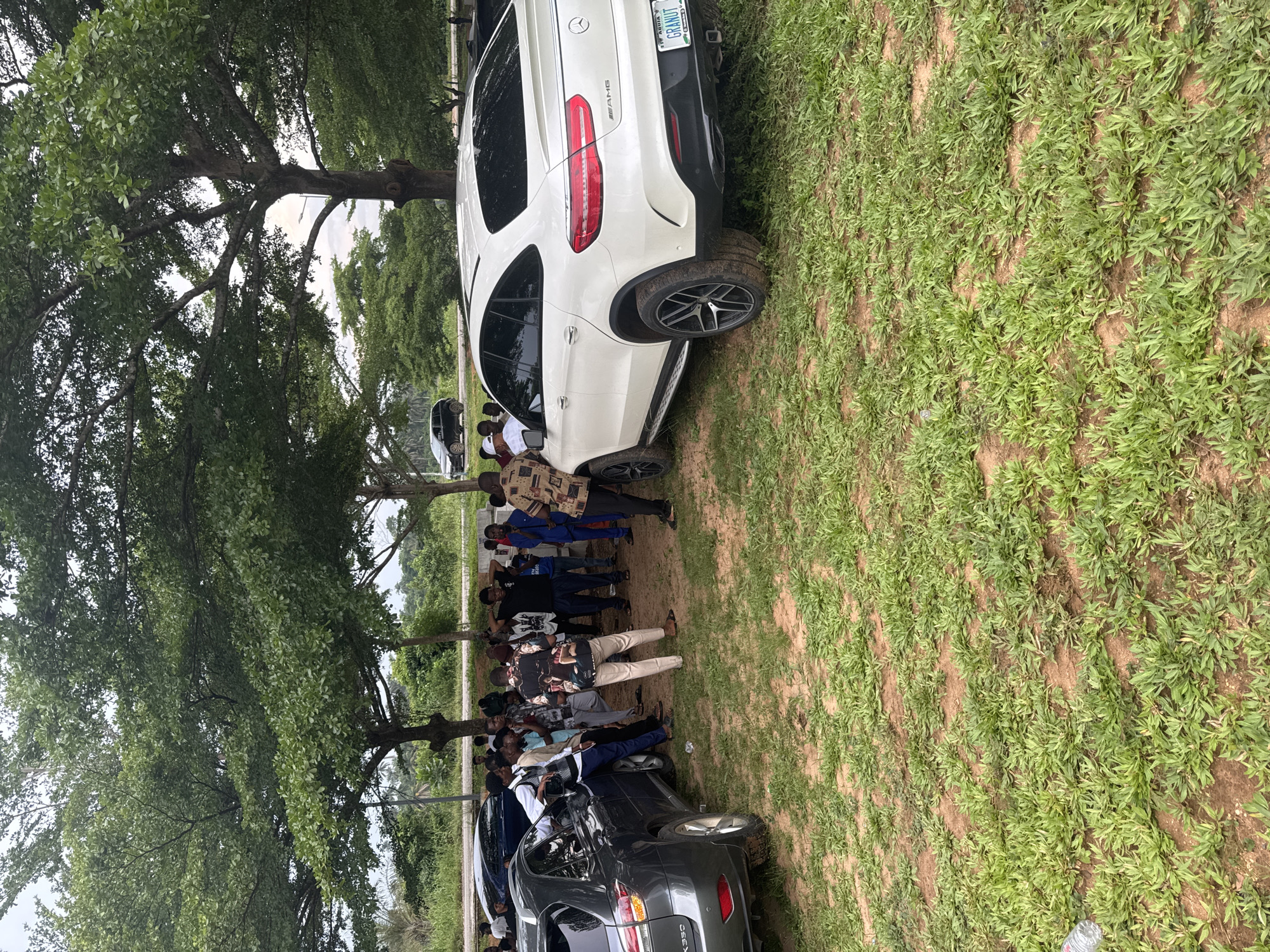 A group of people stand in a grassy area beneath a large tree in Ihiagwa, Nigeria. Two cars, a white SUV and a dark sedan, are parked nearby. The white SUV has a license plate that appears to read "GRAHUT, AGUATA." The ground is a mix of grass and bare earth.
