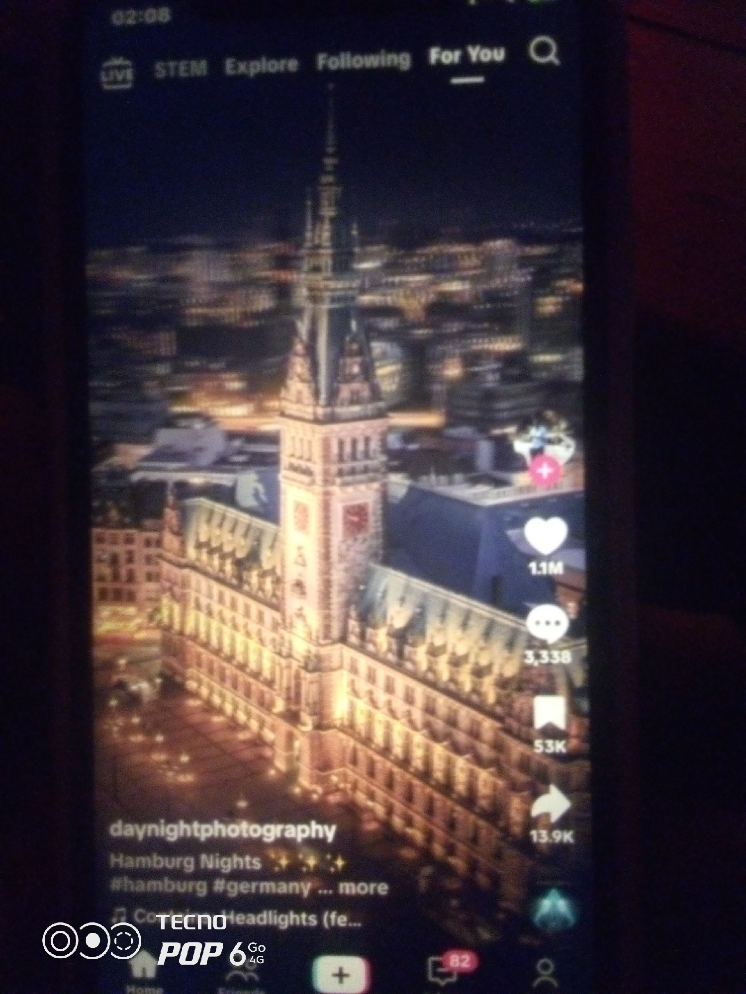 This media file is a vertical video presented on a smartphone screen, likely a social media application. The primary subject is a nighttime cityscape dominated by a large, ornate building with a prominent clock tower, illuminated by warm lights. The surrounding area is also lit, suggesting an urban environment at night.
The video appears to be a visual presentation of a location, possibly a travel or photography post, given the text overlays. The timestamp at the top left indicates "02:08", and the navigation bar at the top shows options like "LIVE", "STEM", "Explore", "Following", and "For You", alongside a search icon.
On the right side of the screen, interaction metrics are visible: a heart icon with "1.1M" (likely likes), a speech bubble icon with "3,338" (comments), a bookmark icon with "53K" (saves), and a share icon with "13.9K" (shares).
Below the main visual, text overlays provide context. The username appears to be "daynightphotography". A caption reads "Hamburg Nights" followed by shimmering emojis, and hashtags like "#hamburg" and "#germany". Another line of text mentions "COTECNO Headlights (fe...".
The bottom of the screen shows a navigation bar with icons for "Home", "Friends", and a plus sign for creating content. The phone model "TECNO POP 6 Go 4G" is also visible, indicating the device on which the video is being viewed. The overall mood conveyed by the nighttime city lights and the engagement metrics suggests a popular and visually appealing content piece. The location context of "Howayi, Nigeria" is relevant to the user viewing or posting this content on their device, but not directly depicted in the video itself.