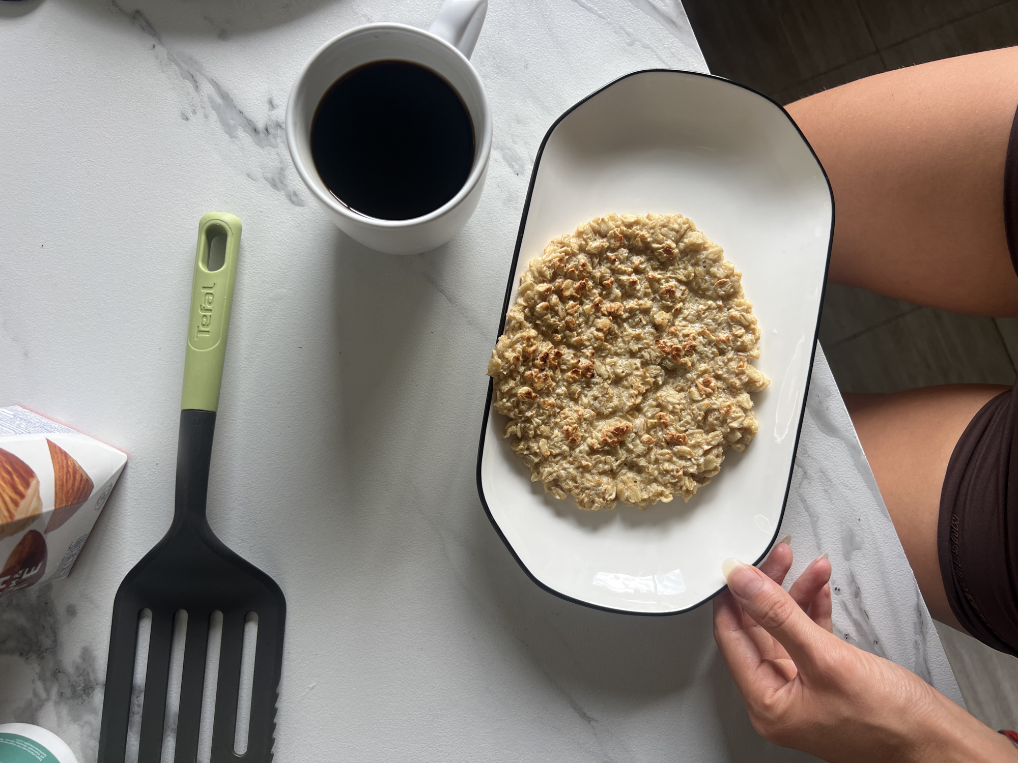 An overhead shot reveals a simple meal setting on a white, marble-patterned tabletop. A person's hands and upper thighs are visible on the right side of the frame, with one hand gently holding a white rectangular plate. The plate, adorned with a black rim, holds a circular portion of baked or pan-fried oatmeal. To the upper left of the plate, a white ceramic mug containing dark black coffee is positioned. Further left, a black slotted spatula with a light green handle lies on the table, featuring the brand name "Tefal" embossed on its handle. In the bottom left corner, a white carton box with visible almonds and the word "Almond" suggests a dairy alternative, and a small, green-lidded container is partially visible beneath it. The scene depicts a quiet indoor moment, likely a breakfast or light meal during the daytime.