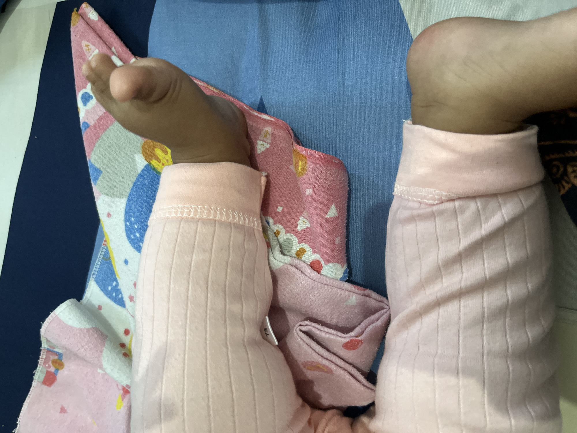 This media file captures a close-up, high-angle view of the lower legs and feet of a young child, likely a baby or toddler, in Lhokseumawe, Indonesia. The child's legs are clad in soft, light pink, vertically ribbed sleepwear or pajamas. The cuffs of the sleepwear appear to be rolled up or gathered around the ankles.
Underneath the child, several layers of fabric are visible. On the left side of the image, there is a pink patterned blanket or towel featuring various colorful shapes including white, blue, and red elements. Below this, a solid blue fabric, possibly a sheet or another blanket, covers a significant portion of the background. On the far right, a dark blue or black fabric with an intricate pattern is partially visible.
The child's bare feet and ankles are seen emerging from the sleepwear. The posture suggests the child is resting or lying down comfortably. A small white tag, possibly from the sleepwear, is visible between the child's left leg and the pink patterned blanket, clearly displaying the number "73". The overall setting appears to be indoors, likely on a bed or a soft surface. No specific time of day or weather conditions are discernible from the image.