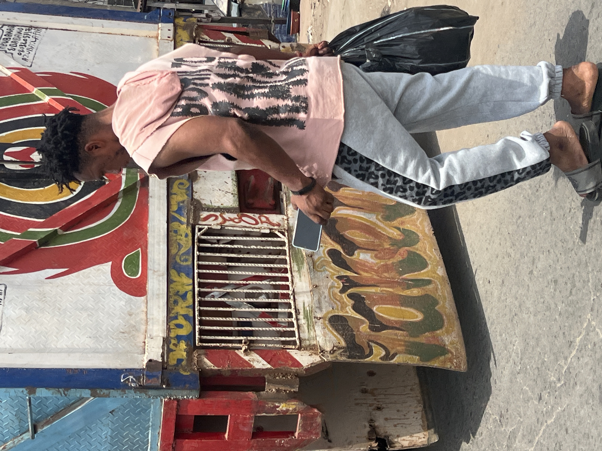 In the city of Onitsha, Nigeria, a man stands next to a brightly painted truck on an overcast day. The man is dressed in a light pink t-shirt with a black and white pattern, light grey sweatpants with black and grey animal print stripe on the side, and grey sandals. He has dark hair and is holding a black cell phone in one hand and a black plastic bag in the other. The truck has multiple colors and shapes painted on the side. The backdrop shows a concrete surface.