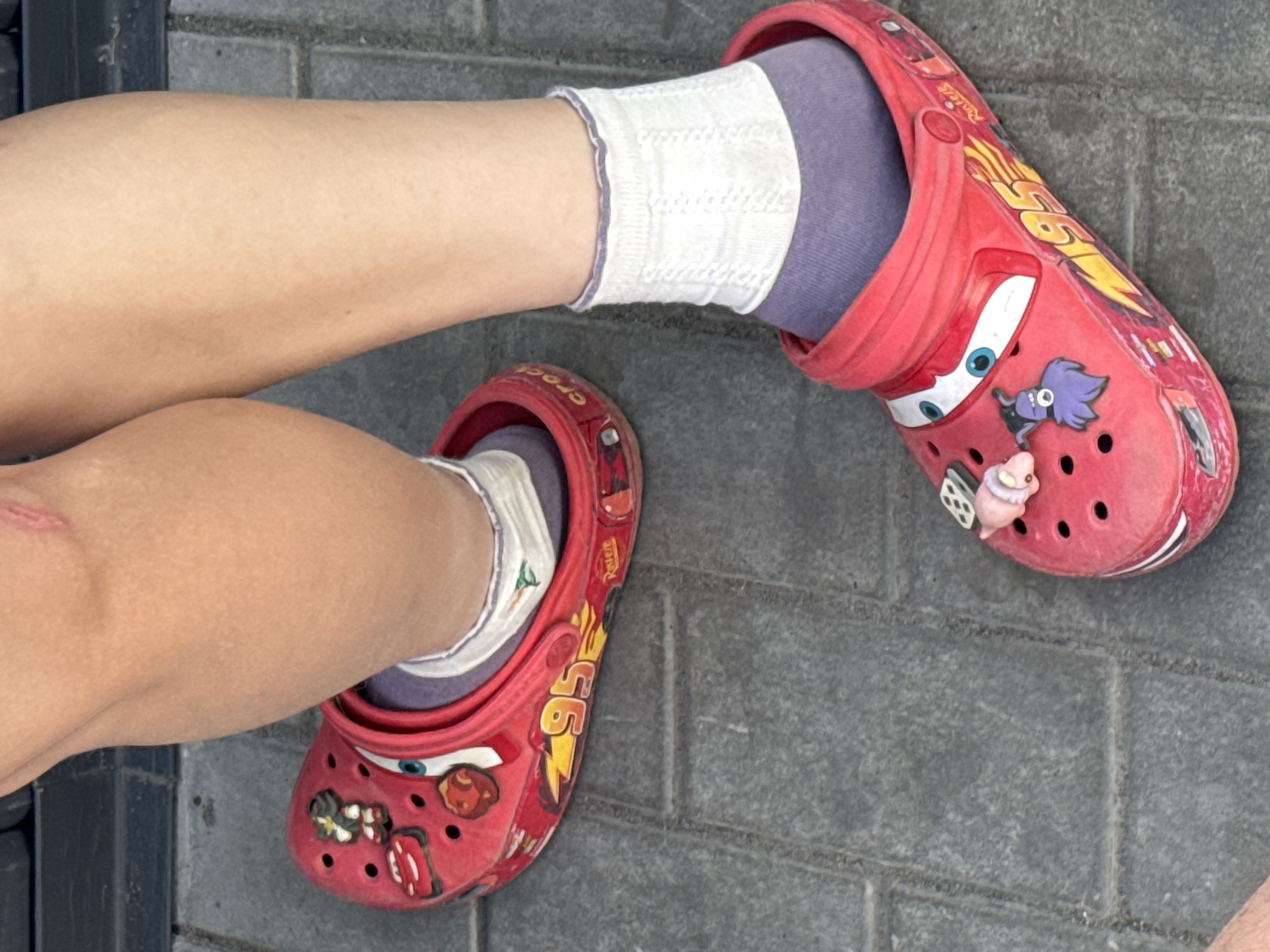 The image shows a person's legs and feet wearing red Crocs with a "Cars" design. The Crocs are adorned with Jibbitz charms, including a purple creature and a pink pig. The person is wearing socks: one foot has a grey sock and the other has a grey sock, both covered by white frilly socks. The feet are placed on a grey brick surface. The location is unknown, and the prompt about Bol'shie Nyrsi, Russia, is not relevant to the visible content.
