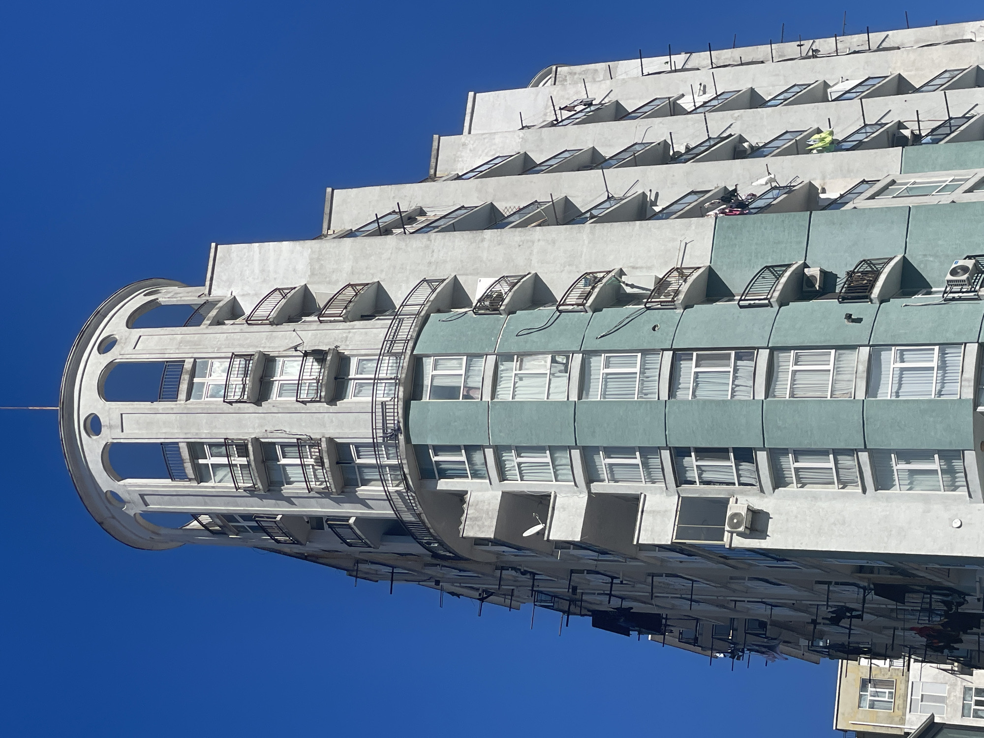 The image captures a portion of a tall, terraced building in Batumi, Georgia, set against a clear blue sky. The building features a distinctive cylindrical section on one side, with multiple arched openings. The facade is predominantly light-colored, with sections painted in a muted teal. Numerous windows line the building's facade. Some upper terraces have small, triangular skylights and metal railings. A series of metal ladders runs vertically along the building. Air conditioning units are visible on some of the floors. Laundry is hung out to dry on the terraces.
