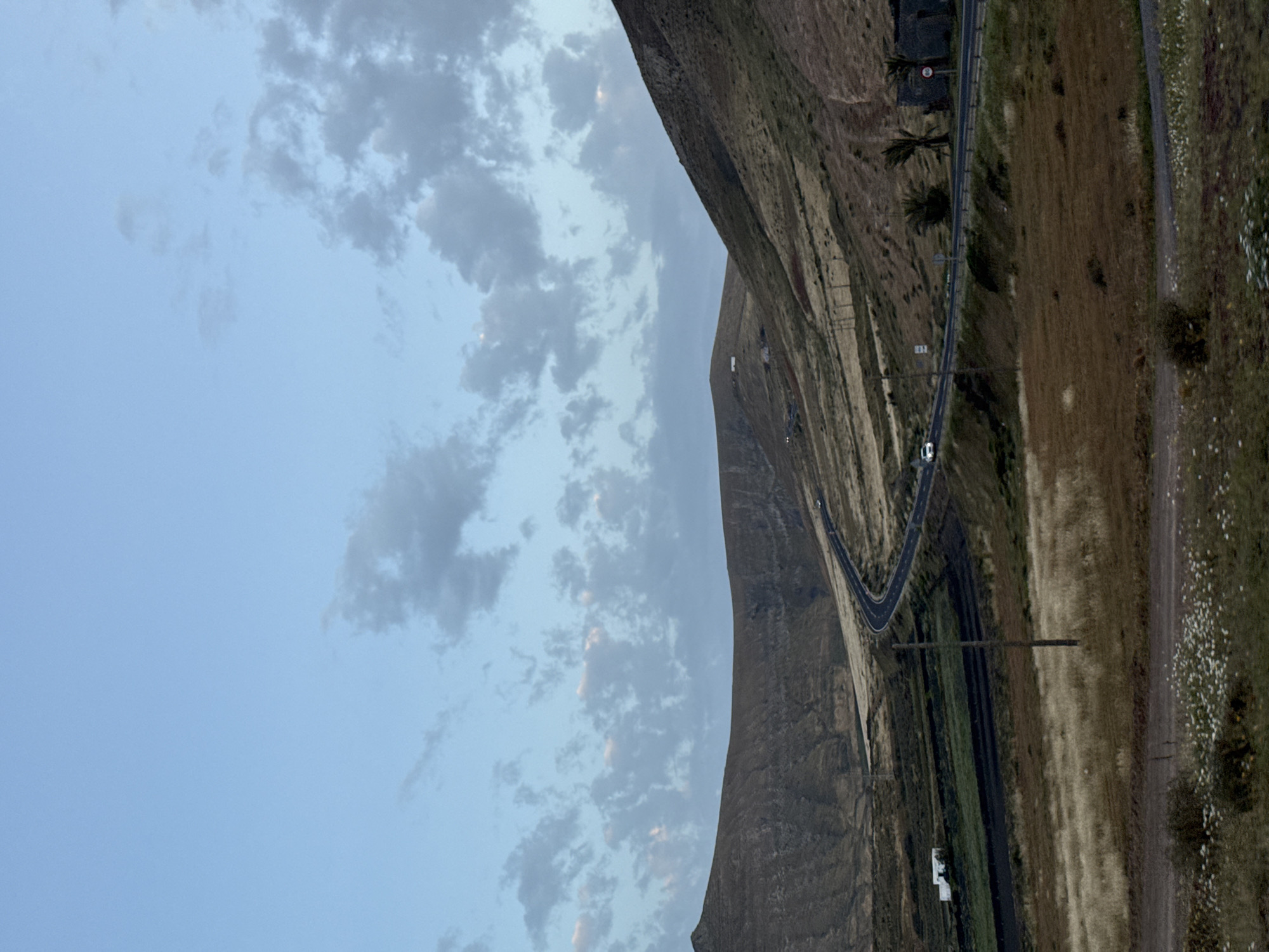 The image shows a landscape in Yaiza, Spain, featuring a winding road adjacent to a steep, rocky mountain range. The sky is partly cloudy with a gradient of light blue. The road is asphalt, with a white vehicle visible. The landscape beside the road varies from dry grass to green vegetation. Utility poles run parallel to the road.