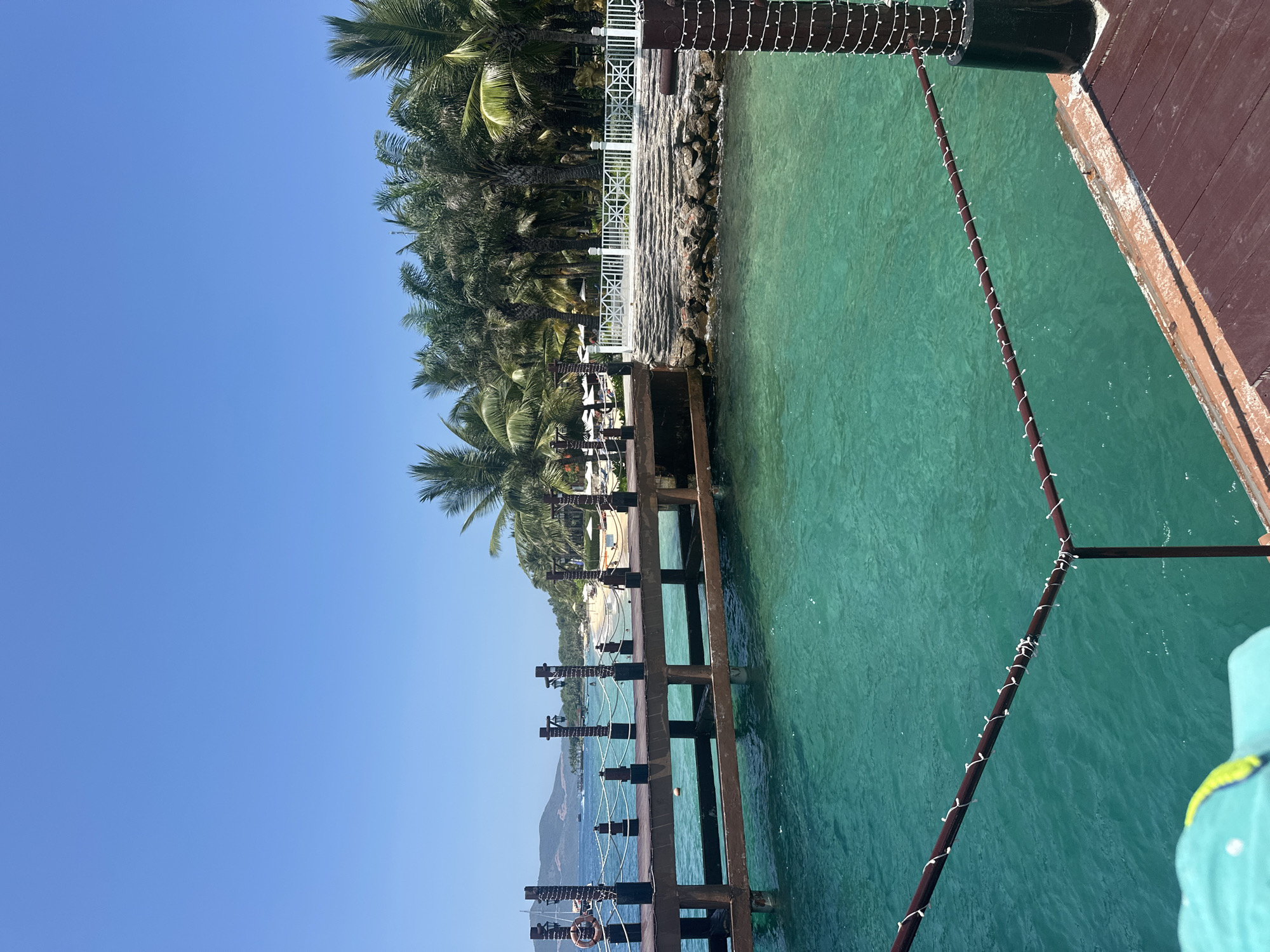 A sunlit view captures a wooden pier extending over turquoise water towards a tropical beach and distant mountains in Nha Trang Ward, Vietnam.
In the immediate foreground, the top of a teal-colored hat with a lime-green brim is partially visible at the bottom left. The dark brown wooden decking of the pier occupies the bottom right, and a horizontal dark brown metal railing, intricately wrapped with white string lights, stretches across the lower portion of the frame. The water below and extending into the midground is a luminous, clear turquoise, exhibiting gentle ripples on its surface.
The central feature is a substantial wooden pier, constructed of dark brown timbers and posts. Each post is embellished with white string lights, and some posts are topped with lanterns. A red-and-white lifebuoy is secured to a post on the left side of the pier. Rope railings connect the pier posts, forming a boundary. To the right of the pier, a rocky embankment rises to a white decorative fence. Behind this fence, a dense grove of palm trees forms a lush green backdrop, with several golden, sculpted figures partially visible among the trees. To the left of the pier, a sandy beach stretches into the distance, populated with multiple white beach umbrellas and sun loungers. Several small, indistinct human figures are discernible on the beach. Beyond the beach, in the deeper blue water, various boats, including sailboats, are scattered.
The background reveals a chain of verdant, vegetated mountains rising across the horizon. Above, the sky is uniformly clear and light blue, completely devoid of clouds.