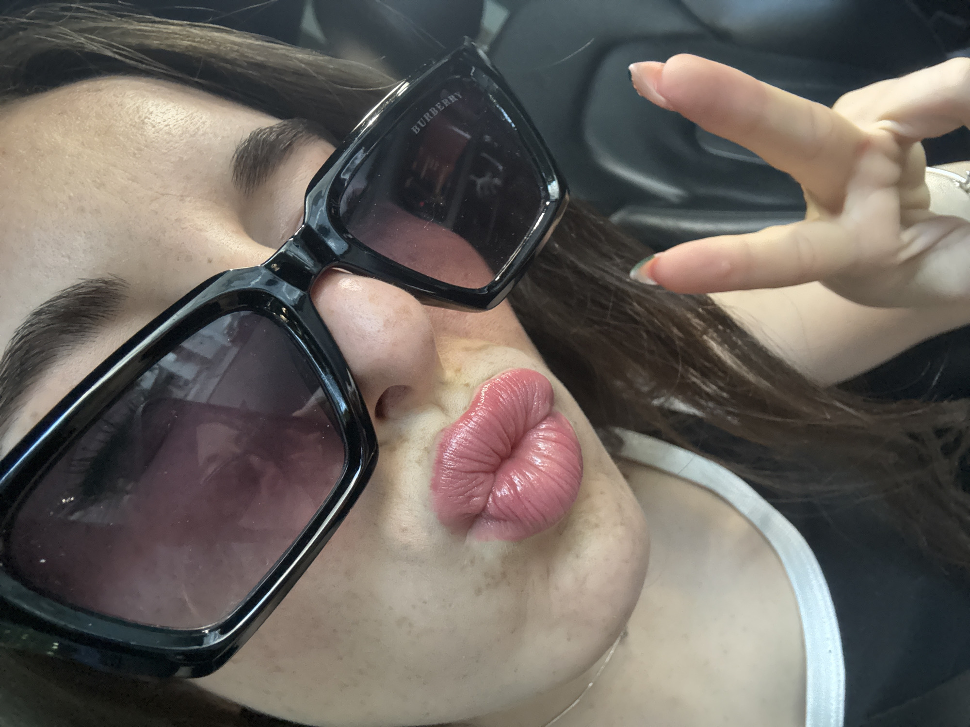 A young woman is depicted in a close-up selfie taken indoors, likely within a vehicle. She wears oversized, black-framed Burberry sunglasses. Her lips are pursed in a kissing gesture. Her right hand is raised, making a "peace" or "victory" sign. She has freckles and a white top with a thin strap. The background is dark, suggesting the interior of a car. There is no visible information to ascertain the location in Plovdiv, Bulgaria.
