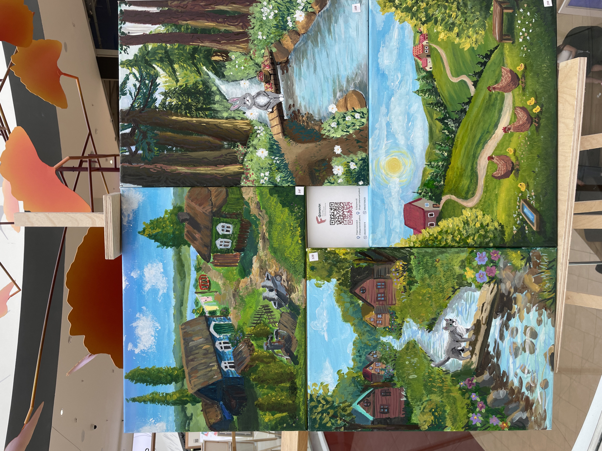 A display of four paintings is shown. The paintings are arranged in a 2x2 grid. The top left painting depicts a village scene with houses, a body of water, and trees under a blue sky with clouds. The top right painting shows a forest scene with a stream, a bridge, and a rabbit. The bottom left painting features a stream flowing through a village with houses and a cat standing on a log. The bottom right painting illustrates a rural landscape with rolling hills, houses, chickens, and chicks. A QR code and some text are visible on the bottom right corner of the top right painting, and a label with "12B" is attached to the top right painting. Another label with "14Г" is attached to the top left painting. A label with "298" is on the bottom right painting, and a label with "18B" is on the bottom right. The paintings are displayed on easels in what appears to be an indoor setting, possibly a gallery or exhibition space. Above the paintings, abstract orange and pink decorative elements are suspended from the ceiling. The lighting suggests an indoor environment. There is no specific information about the city of Perm, Russia, in the image.