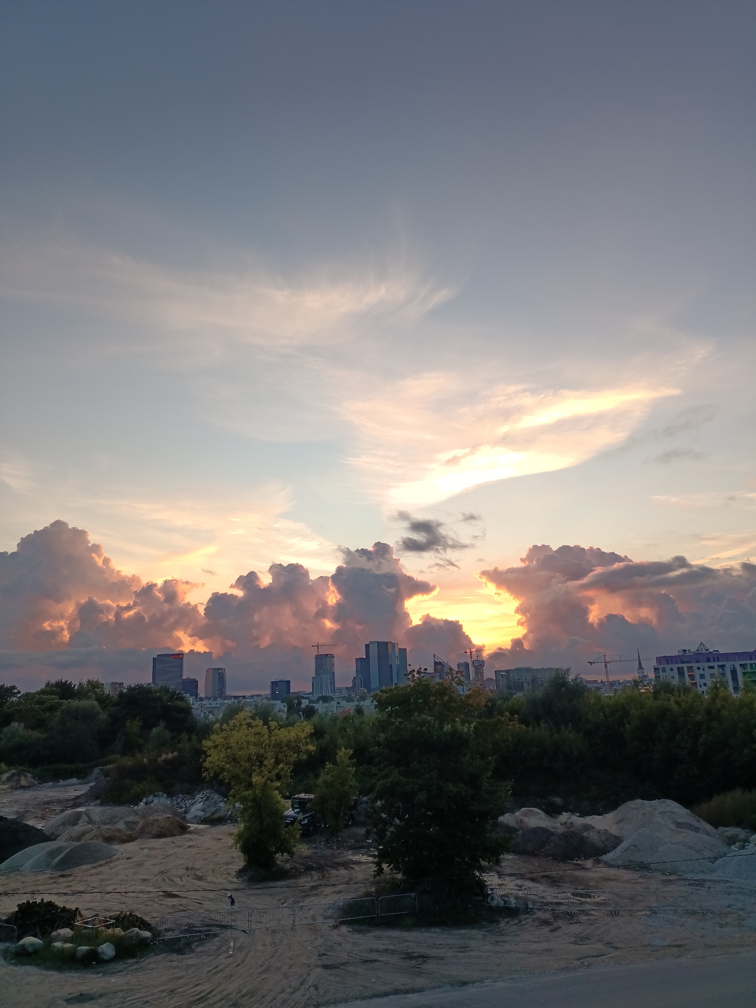 This media file captures a dramatic sunset over the city of Tallinn, Estonia. The sky is filled with large, billowing clouds tinged with vibrant hues of orange, pink, and yellow, indicating the sun is low on the horizon. Wispy cirrus clouds streak across the upper portion of the sky, catching the fading sunlight.
In the mid-ground, a cluster of modern skyscrapers forms the city's skyline. Some buildings appear to be made of glass, reflecting the warm tones of the sunset. Construction cranes are visible among the buildings, suggesting ongoing development in the city.
The foreground depicts a more rugged, undeveloped landscape. Piles of gravel and sand are scattered across a dirt ground, indicative of a construction site or quarry. Several trees, some with green foliage and others with yellowing leaves, dot the landscape, suggesting it is either late summer or early autumn. A dark-colored utility vehicle is parked amidst the trees. Metal barricades and some bundled construction materials are also present in the foreground.
The overall mood is one of natural beauty and urban development coexisting. The vibrant sunset provides a striking backdrop to the emerging city, creating a sense of awe and anticipation. The time of day is clearly evening, as evidenced by the sunset colors and the low angle of the sun. No visible text is present in the image.