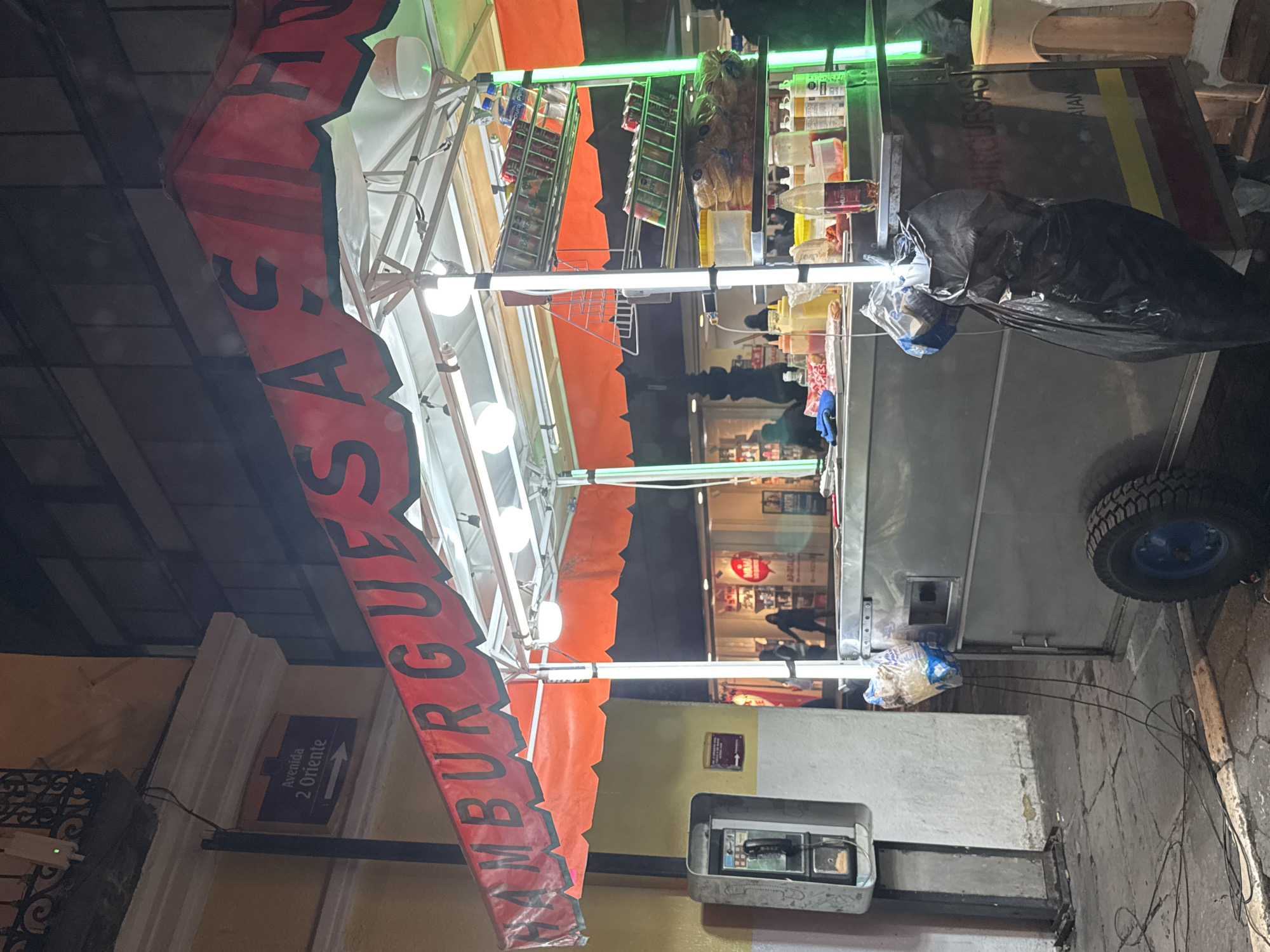 A nighttime scene in Puebla, Mexico shows a roadside "Hamburguesa" food cart. The cart, illuminated by fluorescent green lights and white bulbs, is positioned on a street near a blue street sign that reads "Avenida 2 Oriente" with a right-pointing arrow. An old payphone booth stands near the cart. The cart's side faces the camera, revealing its contents, including shelves of beverages and condiments. A large black trash bag is draped over the cart's side, and a wheel is visible. Several figures can be seen walking in the background.