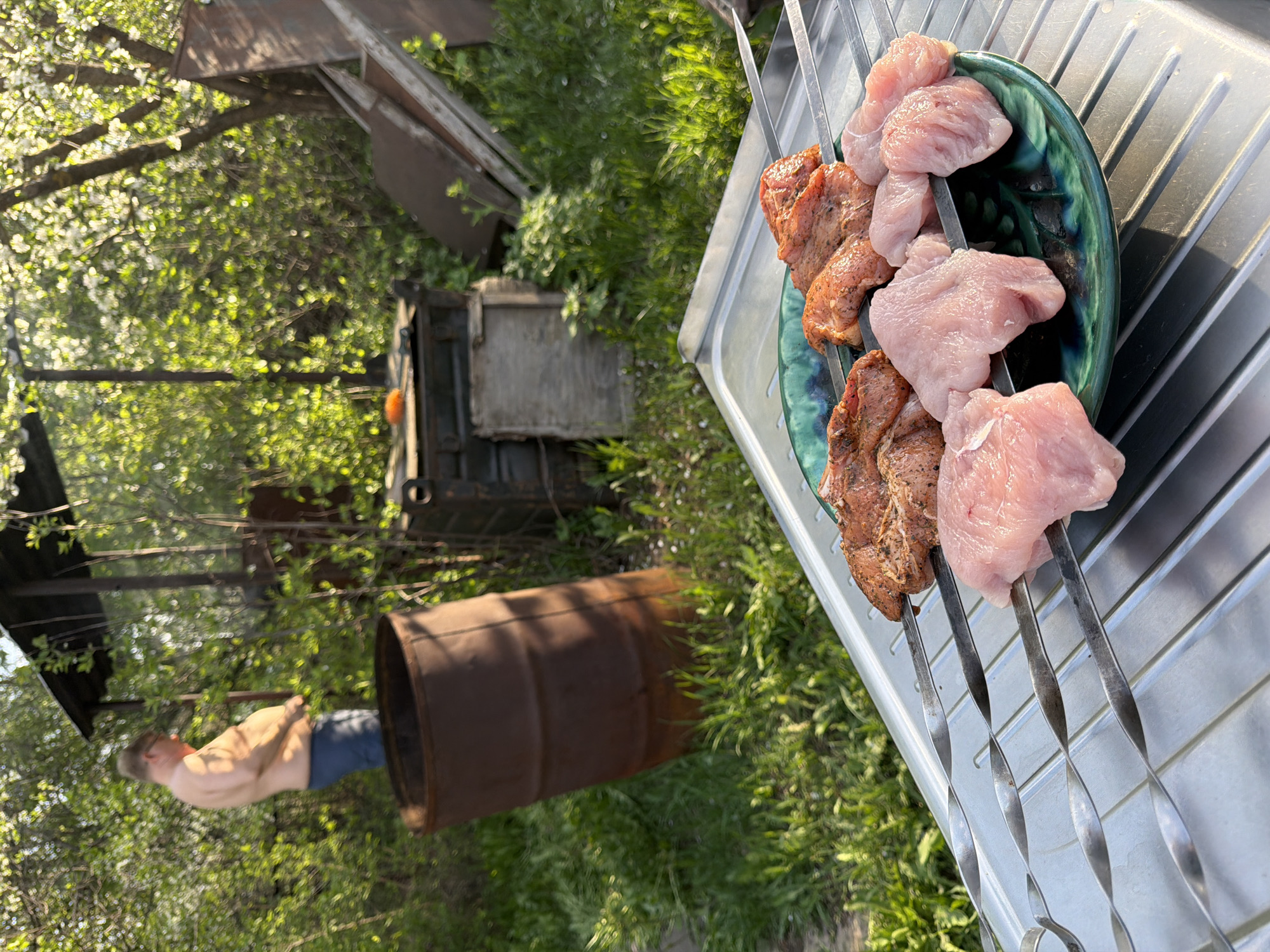 **Scene Description**
The scene is an outdoor setting in Izhevsk, Russia, featuring several elements. In the foreground are metal skewers loaded with raw meat, both marinated pork and what appears to be chicken, resting on a green plate and a silvery metal surface, likely a grill.
In the mid-ground, a bare-chested man wearing blue shorts is standing next to a rusty metal barrel. There's a wooden structure and dense greenery, including trees with white blossoms, in the background.
An old, weathered wooden box or small shed is also present. The overall environment suggests a backyard or an outdoor cooking area.