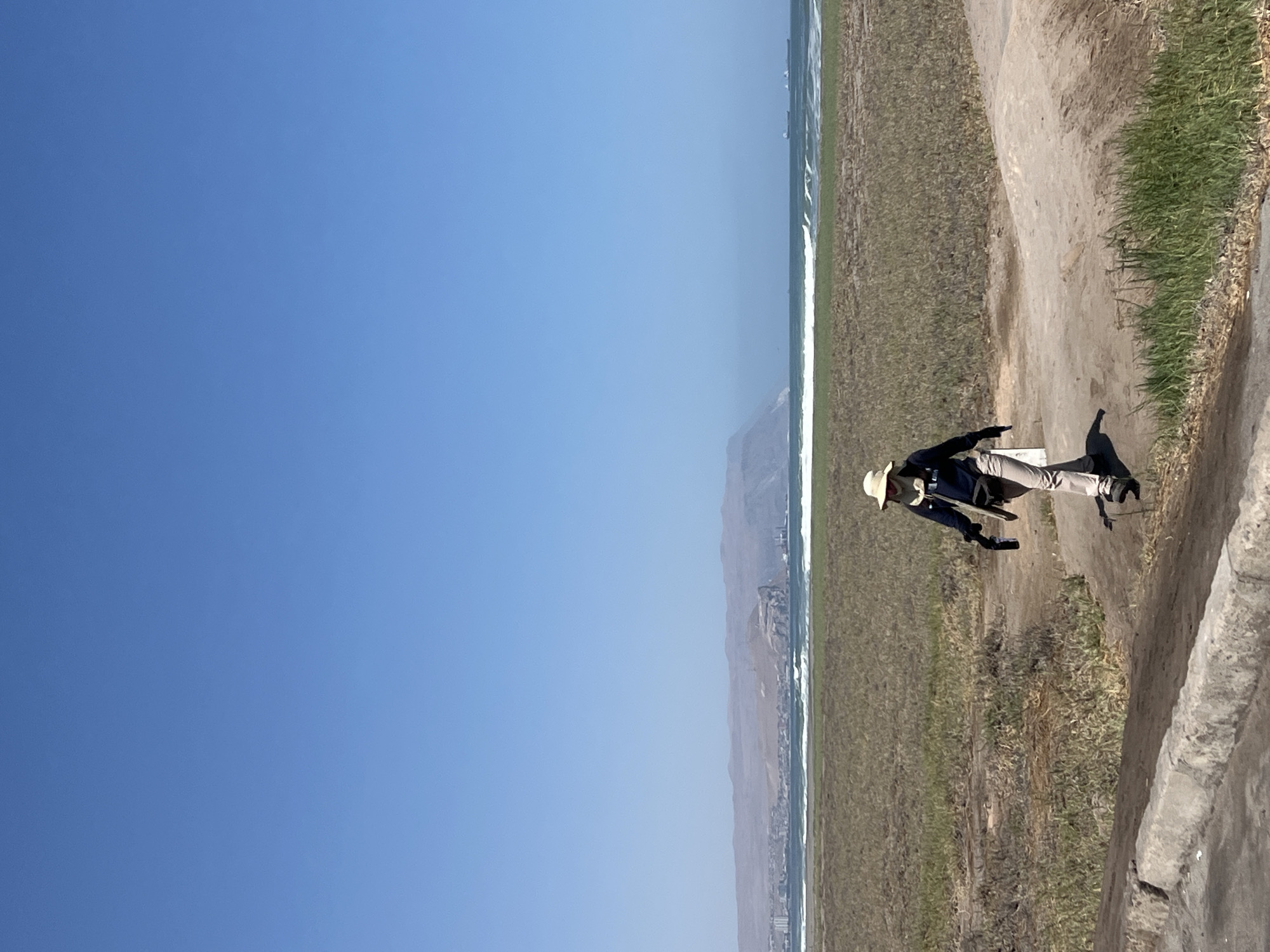 A landscape shot in Arica, Chile, oriented vertically, showcases a vibrant blue sky dominating the left side, transitioning to a scene on the right. The Pacific Ocean, with subtle waves, borders a stretch of arid land. A mountain range is partially visible in the distance.
On a dirt path, a person stands wearing a straw hat, a dark jacket, light-colored pants, and dark footwear. A shadow is cast next to them on the path. A grassy area extends beside the path towards the ocean. A low, gray concrete structure lines the edge of the path, providing a boundary.