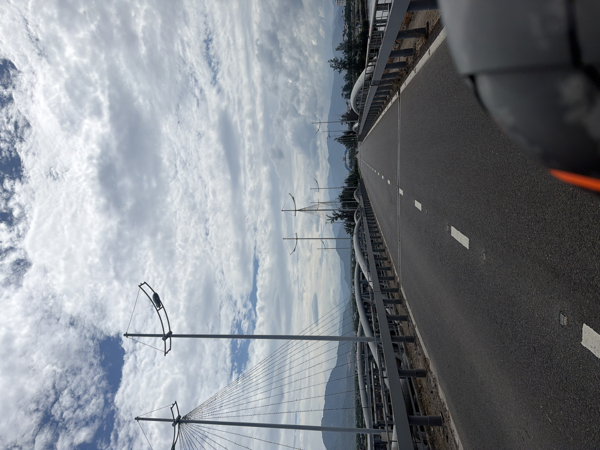 The image presents a daytime, eye-level perspective along an empty bridge or elevated roadway in Bac Cam Ranh Ward, Vietnam. The scene is dominated by a dark asphalt road surface stretching into the distance on the right, bordered by a metal guardrail on the left. White dashed lines mark the lanes, with additional reflective markers embedded in the road. In the upper right corner, the dark gray/black bodywork and an orange reflector of a vehicle are partially visible, suggesting the photo was taken from a car or motorcycle.
To the left of the road, distinctive gray streetlights or decorative poles with arching tops and radiating wires or cables extend upwards and outwards, creating a striking visual element against the sky. These structures feature a black, possibly bird-like, ornament on one of the arches. Beyond the immediate bridge structures, a hazy mountain range forms the backdrop, disappearing into the atmospheric distance.
The sky is largely overcast with extensive white and light gray stratocumulus clouds, though patches of clear blue sky are visible, particularly towards the bottom left. The overall lighting suggests a bright but diffused daylight, possibly late morning or early afternoon. No people, other vehicles, or discernable text are visible in the image. The atmosphere is calm and serene.