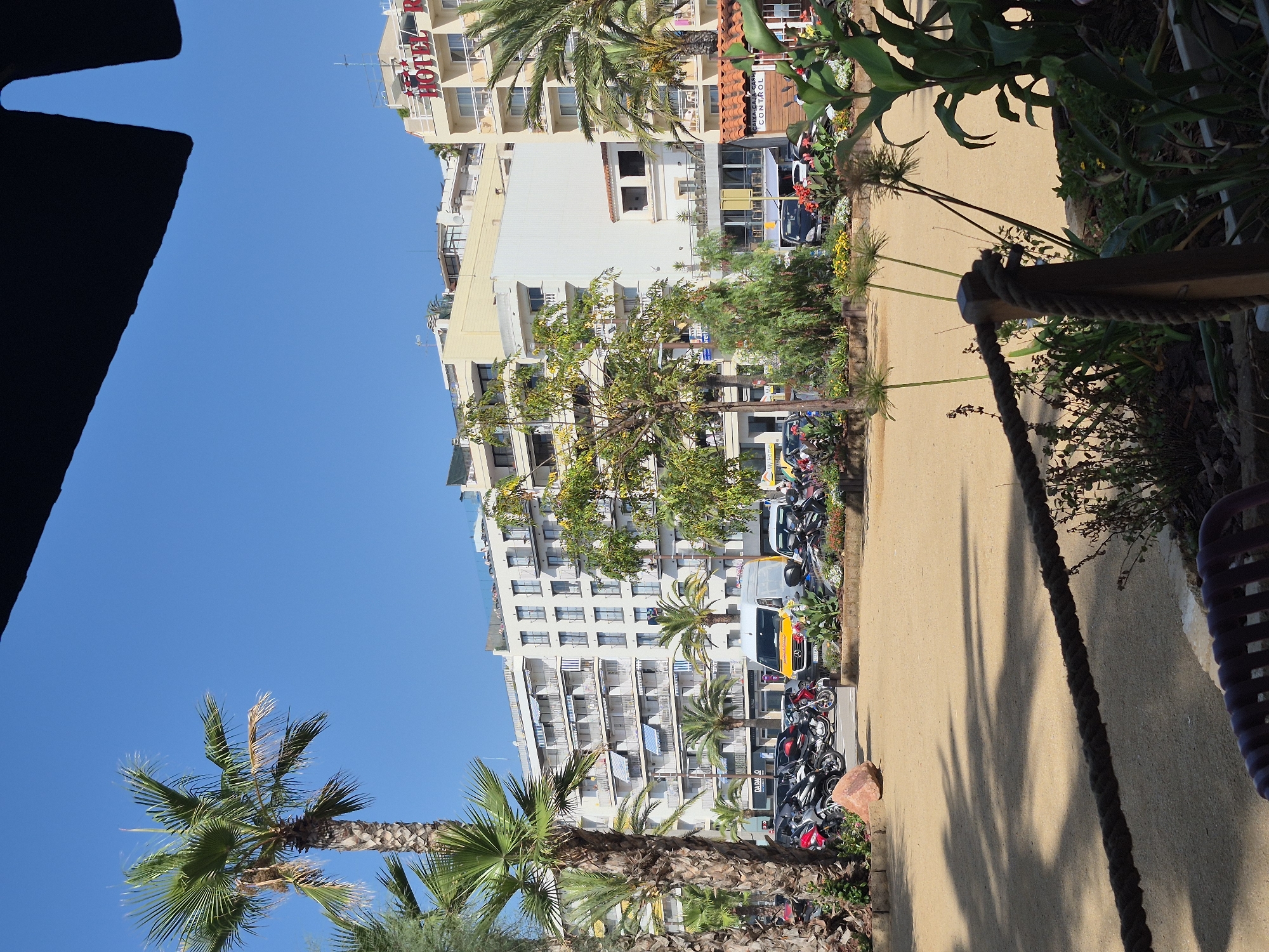 This image depicts a street scene in Lloret de Mar, Spain, on a sunny day. The foreground features a sandy area with a thick rope barrier. In the mid-ground, there is a parking area with several motorcycles and a white van. Beyond the parking area, a row of apartment buildings and a hotel with red lettering visible on its facade line the street. Palm trees and other greenery are scattered throughout the scene, adding to the coastal atmosphere. The sky is clear and blue. There are no visible people in the image.