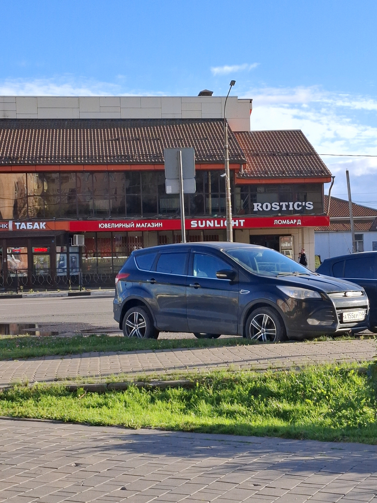 An exterior, eye-level, medium shot captures a dark-colored SUV parked on a paved area next to a grassy strip. The SUV is positioned in the foreground, with its front end slightly angled towards the right. Behind the SUV, a storefront with large glass windows and a red awning is visible. Signs on the storefront read "ТАБАК" (Tobacco), "Ювелирный магазин" (Jewelry store), "SUNLIGHT", and "ЛОМБАРД" (Pawnshop). The architecture of the building features a tiled roof. The sky above is blue with scattered white clouds, and a street light stands to the left of the building. The ground in the foreground is paved with interlocking rectangular stones. A person is partially visible entering the building through a doorway on the right side of the storefront. The license plate of the SUV reads "А 155 EP 750".