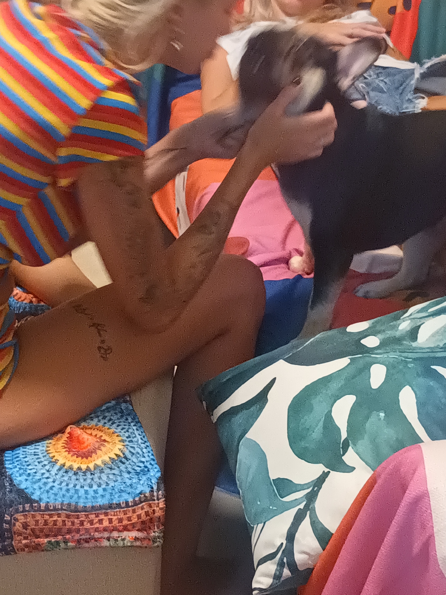 A person with blonde hair is wearing a multi-colored striped shirt and is seated. A person is sitting next to her, wearing an orange shirt with a pink accent. The person is holding a black and white dog with its paws on a blue denim piece. A person's arm and leg are also visible, with a tattoo on the leg. The person is sitting on a colorful pillow. A pillow with a green leaf design is also present in the scene. The location in Ban Nong Khon, Thailand, is not explicitly evident in the image.