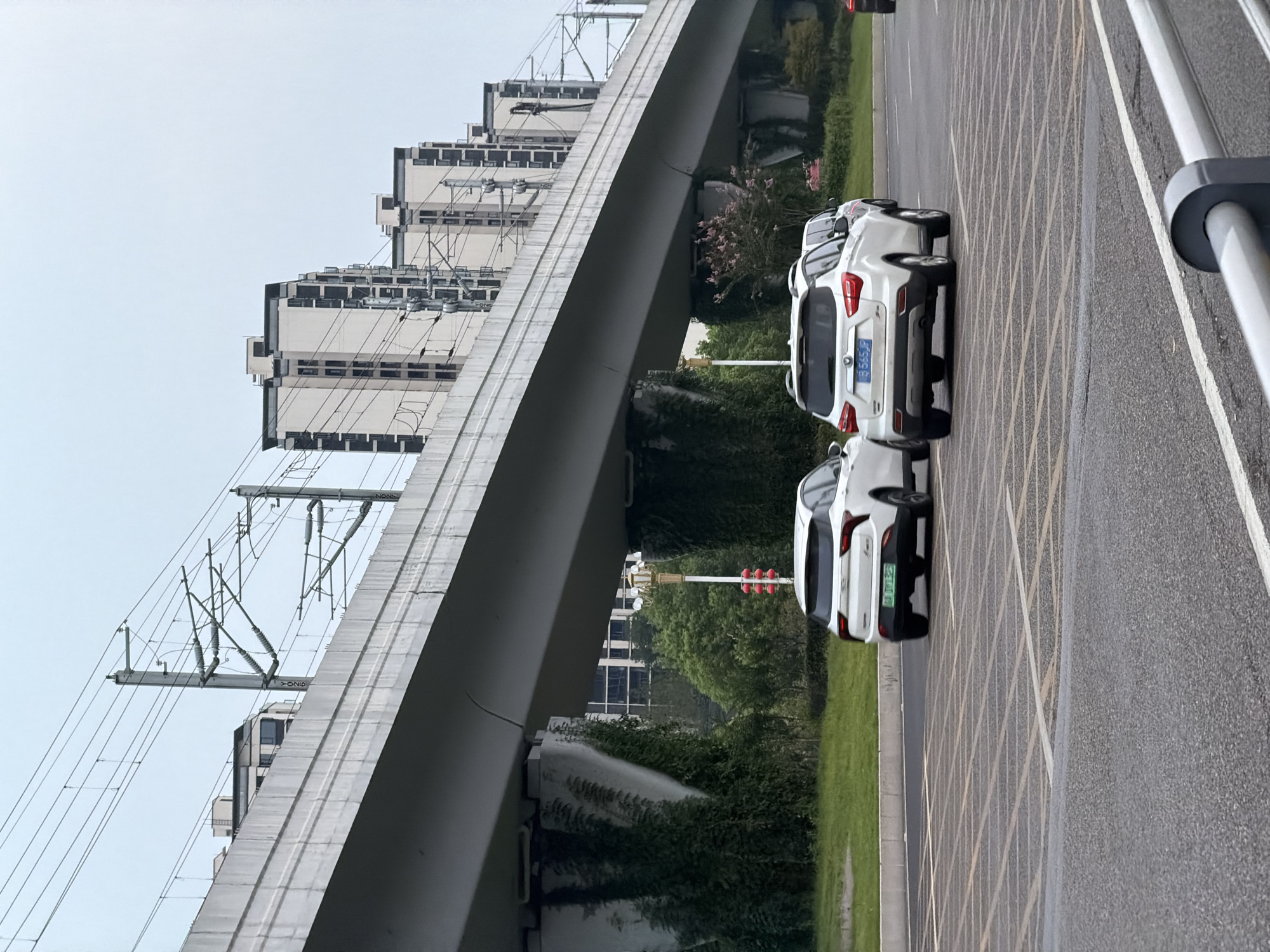 In Jiangjin, China, a white SUV with license plate B 565JP is driving on a road beneath an elevated railway or overpass. The SUV is in the right lane of two visible lanes, with another white SUV to its left and slightly ahead. The overpass is supported by concrete pillars covered in ivy. In the background, several modern, high-rise buildings with a light-colored facade and dark trim are visible, along with overhead power lines for the railway. The sky is overcast. The road surface is asphalt with painted lane markings and a crosswalk pattern.