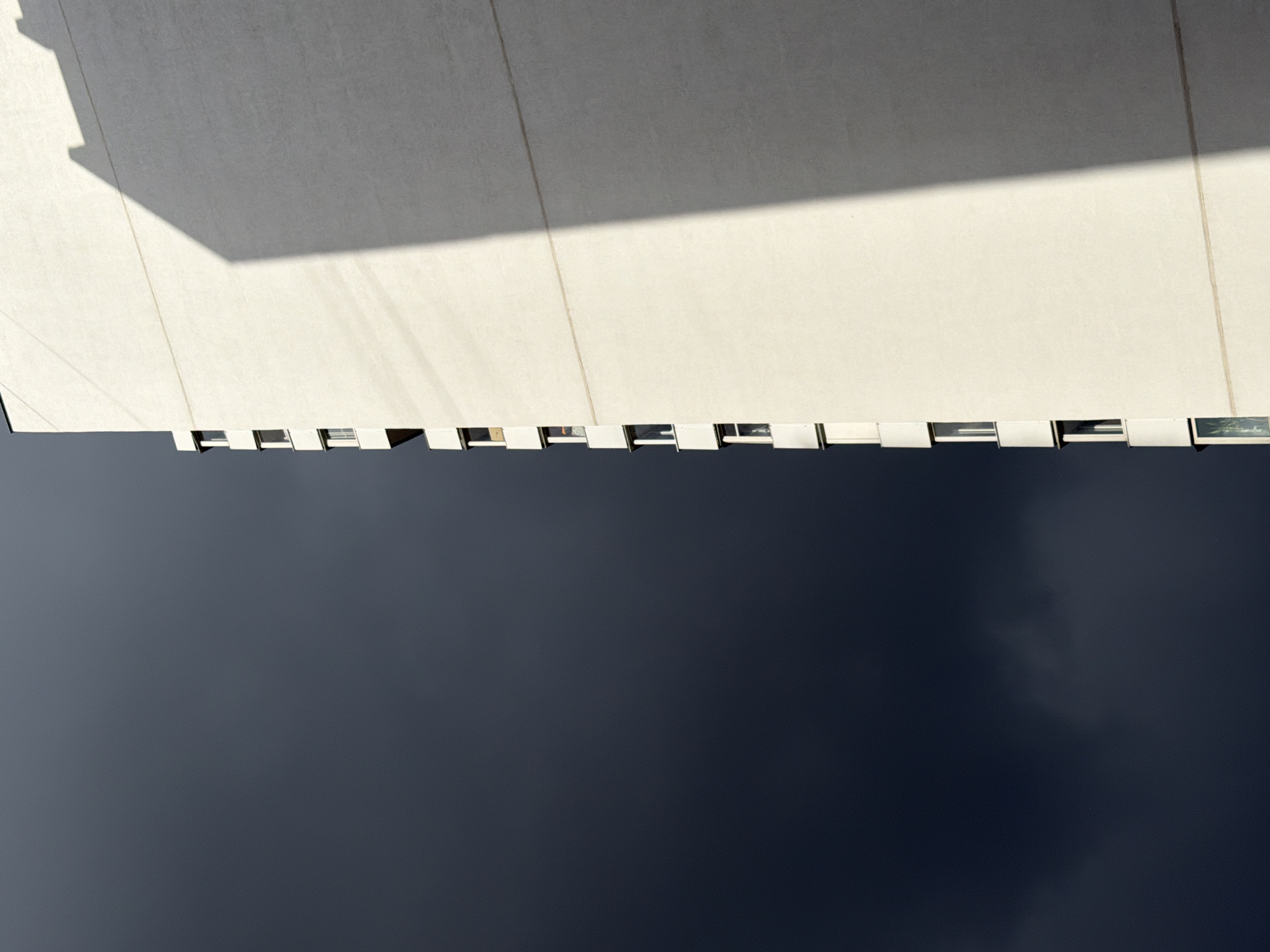 A low-angle, upward shot depicts a section of a building exterior, possibly located in Khimki, Russia, juxtaposed against a dark, clouded sky. The building’s facade appears to be composed of large, rectangular panels, with a light-colored surface partially covered by shadow. Along the lower edge of the building, a series of narrow, rectangular windows are visible, separated by vertical divisions. The sky, occupying the majority of the frame, is a deep, muted blue-gray, showing indistinct, cloudy formations.