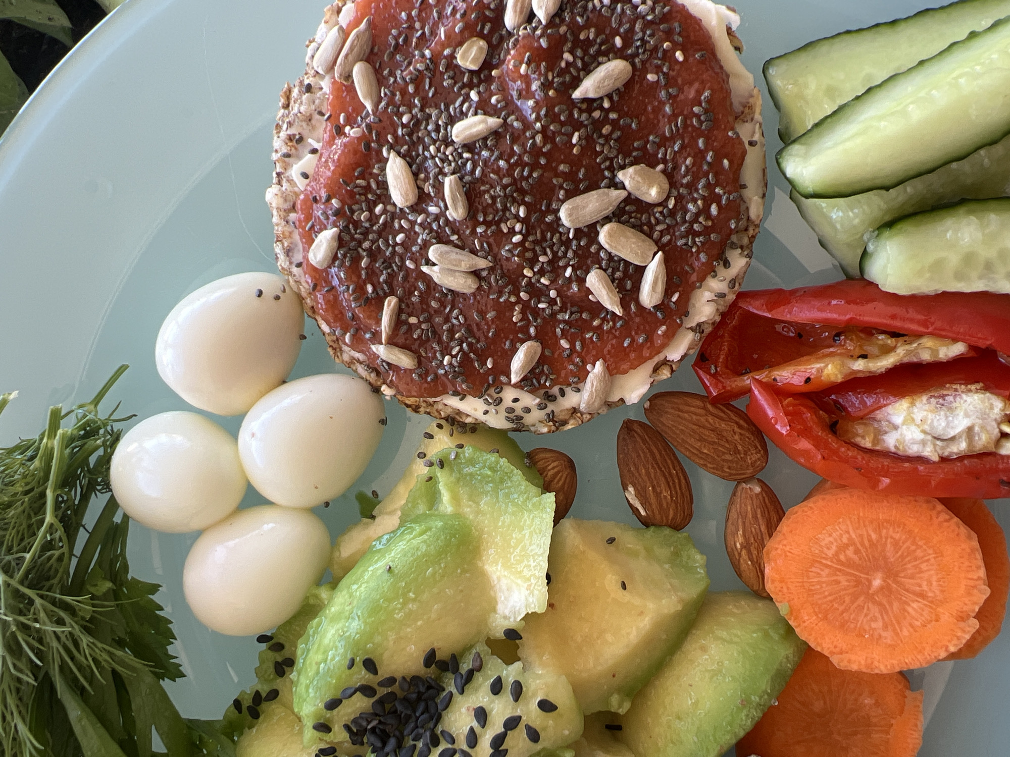A close-up, overhead shot of a light blue plate filled with a healthy meal. The meal includes several sliced pieces of avocado sprinkled with black sesame seeds, four hard-boiled quail eggs, a round crispbread topped with a red spread, sunflower seeds, and chia seeds, a portion of sliced carrots, some almonds, sliced red bell pepper, and sliced cucumbers. A sprig of fresh dill is visible at the top of the plate. The lighting suggests it is daytime. The scene appears to be indoors, likely a kitchen or dining area. There are no people or animals present in the image. The image does not provide any information about its location in Novosibirsk, Russia.