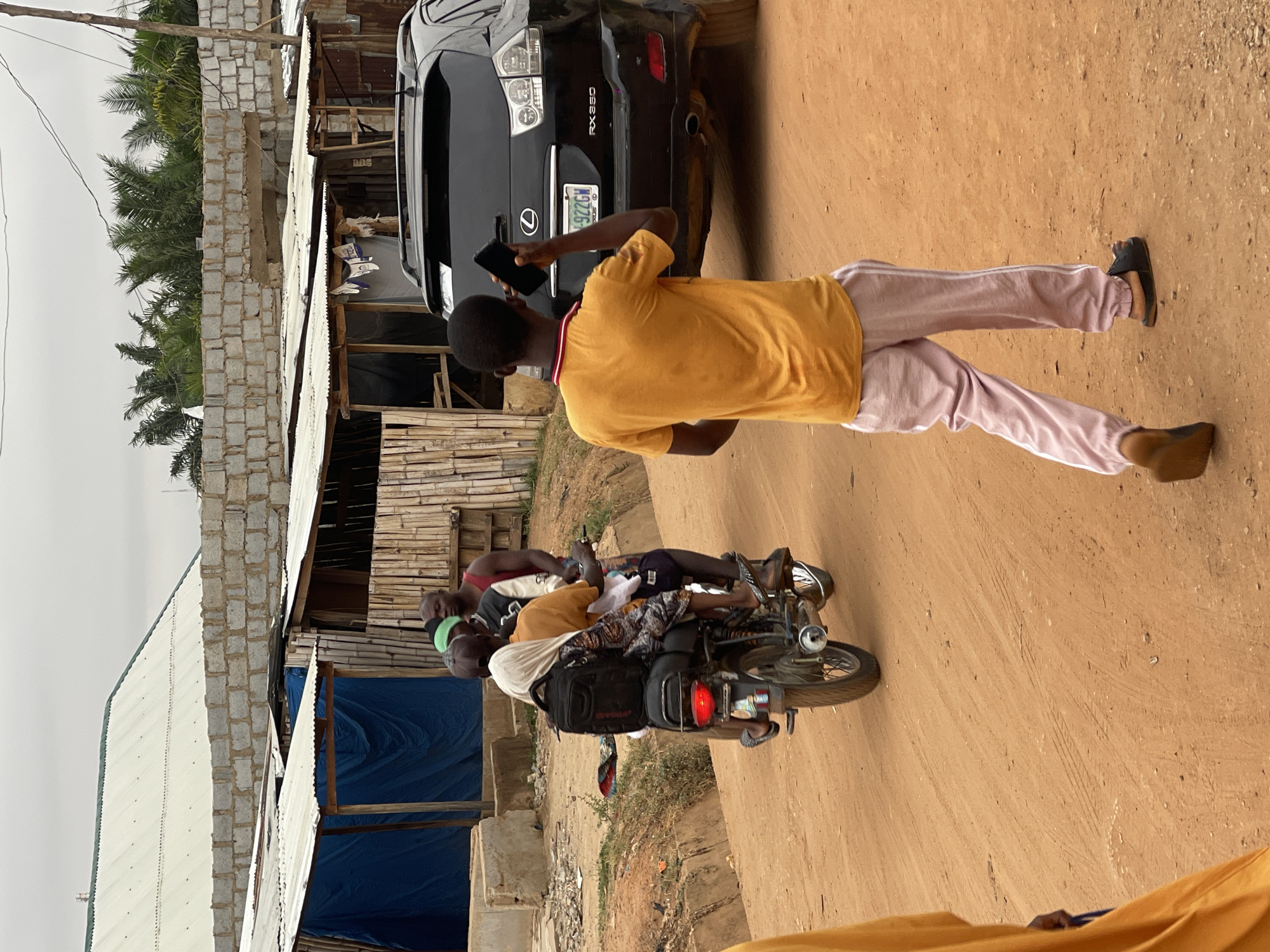 The scene is located in Kuchingoro, Nigeria. A man wearing a yellow shirt and pink pants is standing next to a black RX 350 vehicle, holding a phone in his hand. Nearby, a motorcycle with three people is visible. The environment consists of simple buildings, possibly homes or shops, with various construction materials like corrugated metal and brick. The ground is dirt. The sky is overcast.