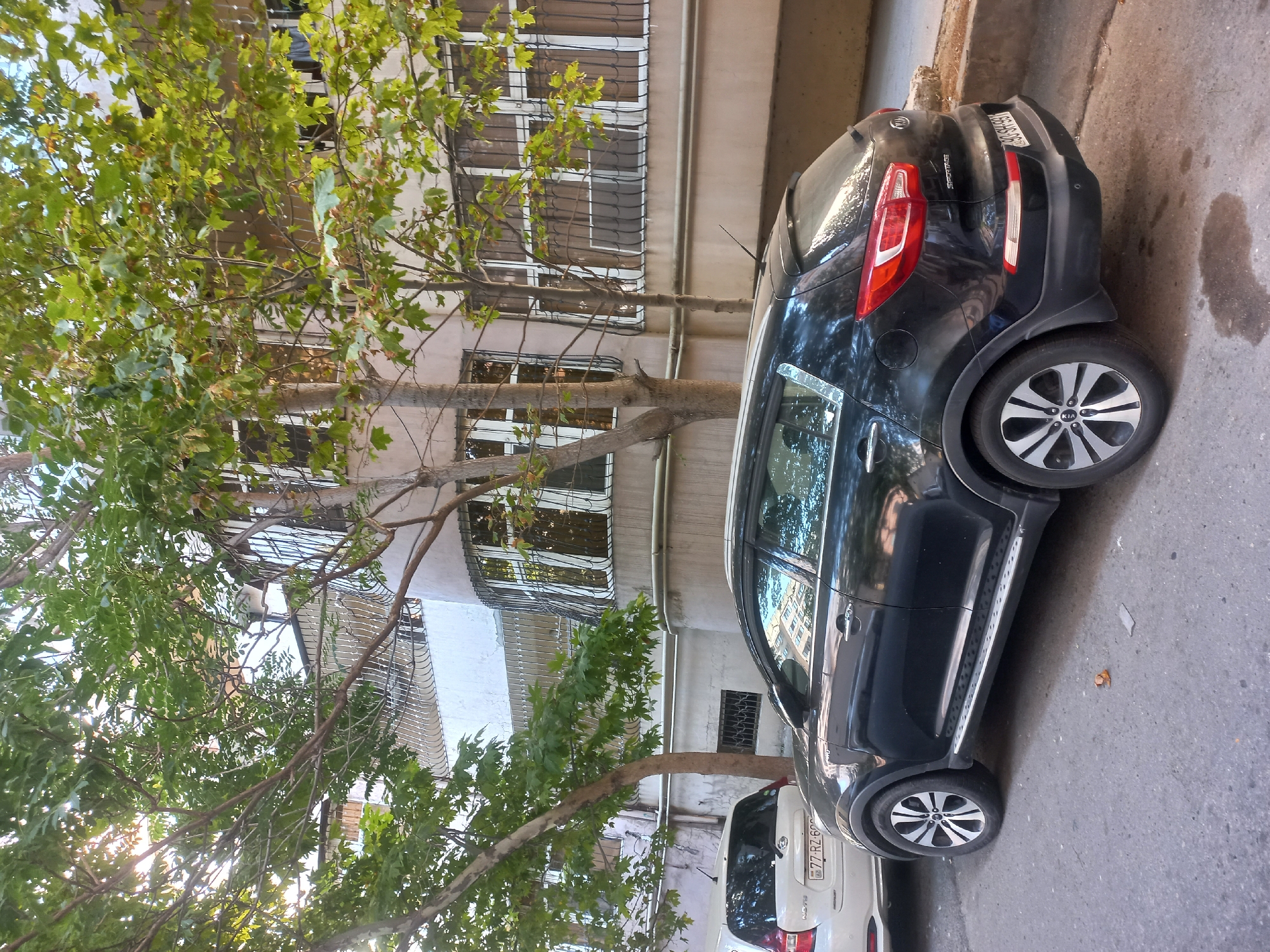In Baku, Azerbaijan, a black Kia Sportage SUV is parked on a paved city street next to a white vehicle. The SUV has tinted windows and is parked in front of an apartment building. The building has multiple windows with metal bars and balconies. Trees with green leaves are in the foreground, casting shadows on the street. The sky is visible in the upper left corner, with sunlight filtering through the trees. A license plate on the white car reads "77-RZ-600."