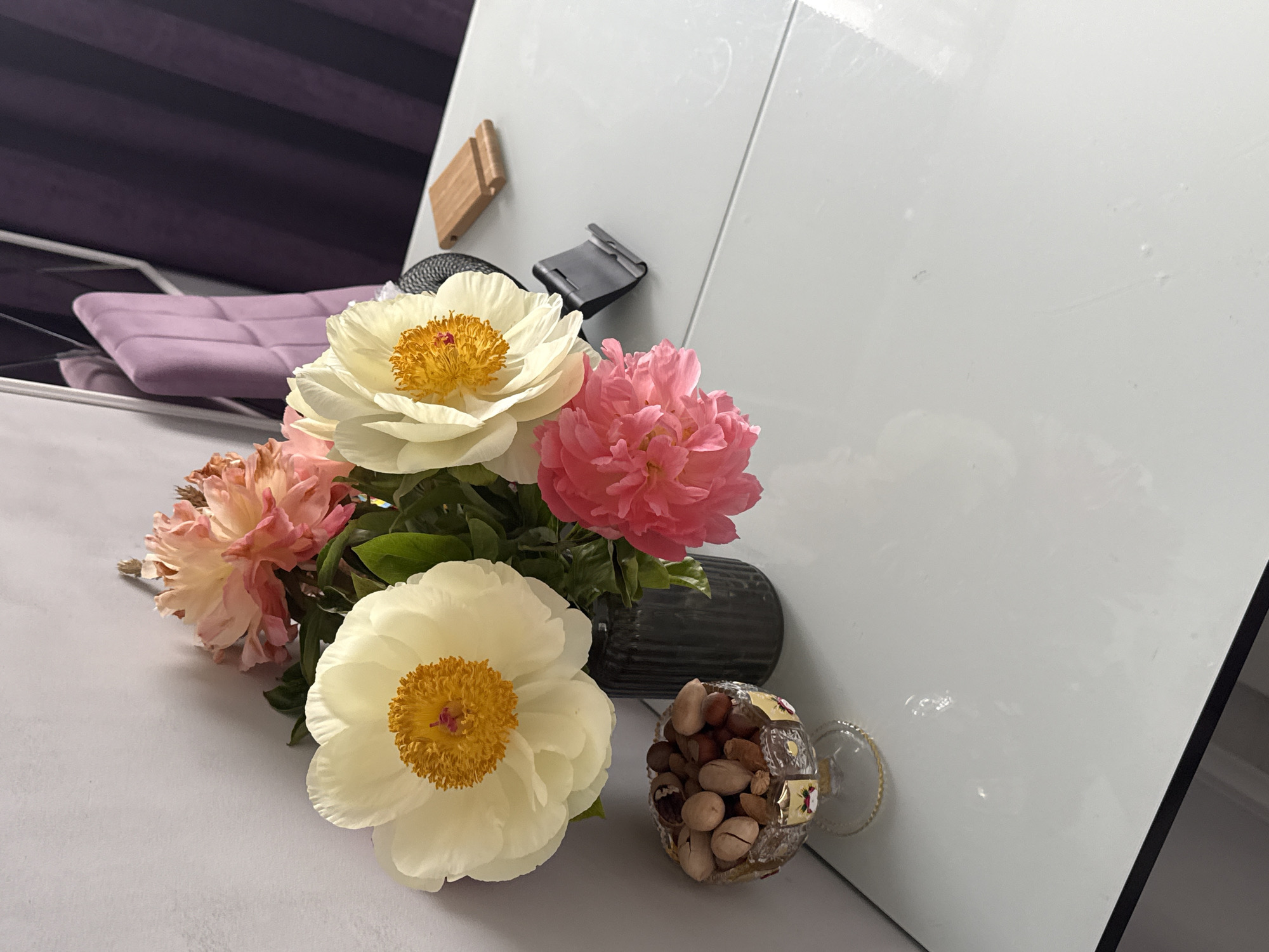 The image presents a bouquet of flowers, predominantly white and pink peonies, arranged in a dark vase alongside a small glass filled with mixed nuts. These items are positioned on a white, reflective surface. The background includes a white panel, possibly a door or furniture piece, adorned with two small, rectangular objects. To the left, a glimpse of a purple cushion and reflective surface is visible. The scene appears to be indoors, with the lack of specific environmental cues preventing determination of the precise location.