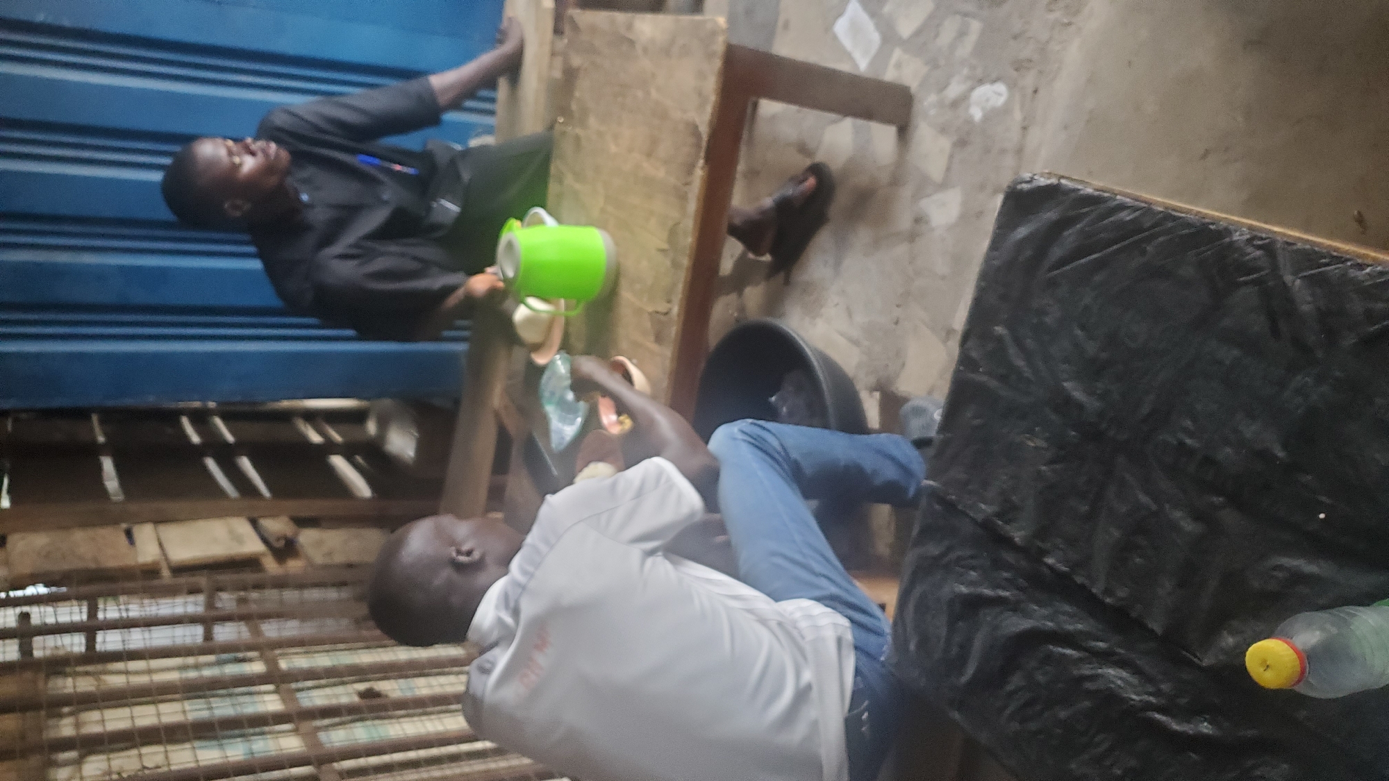 In Jalingo, Nigeria, two men are seated at a wooden table in what appears to be a modest eatery. One man, wearing a white t-shirt and blue jeans, is turned away from the camera, engaged in eating. The other man, in a black shirt and trousers, faces the camera, looking to his right. On the table between them sits a green thermal carafe, plates with food, and a plastic bag. A large black bowl is positioned on the floor near the man in white, and another black bowl is visible behind him. To the left of the frame, a metallic cage or structure is present, and the background features corrugated blue metal sheeting. In the foreground, a portion of a table covered in black plastic is visible, with a plastic bottle and its yellow cap in the lower left corner. The environment suggests a functional, unpretentious setting, possibly an open-air or semi-enclosed food stall.