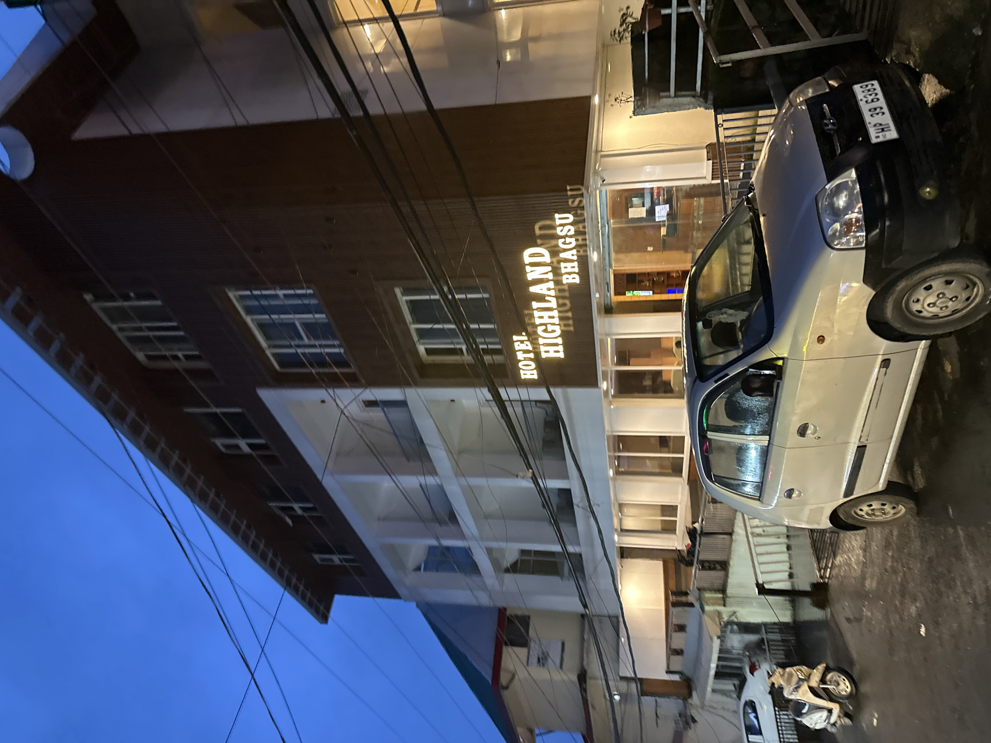 The image shows the exterior of the Hotel Highland Bhagsu. The hotel is a multi-story building with a modern facade, featuring a combination of dark brown and white elements. The name of the hotel is prominently displayed in illuminated signage. In front of the hotel, a silver hatchback car with license plate "HP 39 6389" is parked on a wet, dark asphalt surface, suggesting recent rain. To the left of the car, a white scooter is also parked. The scene is illuminated by streetlights and the hotel's interior lights, creating a twilight ambiance. Overhead power lines are visible, crisscrossing the sky. The overall impression is of a hotel entrance area in a town or city.