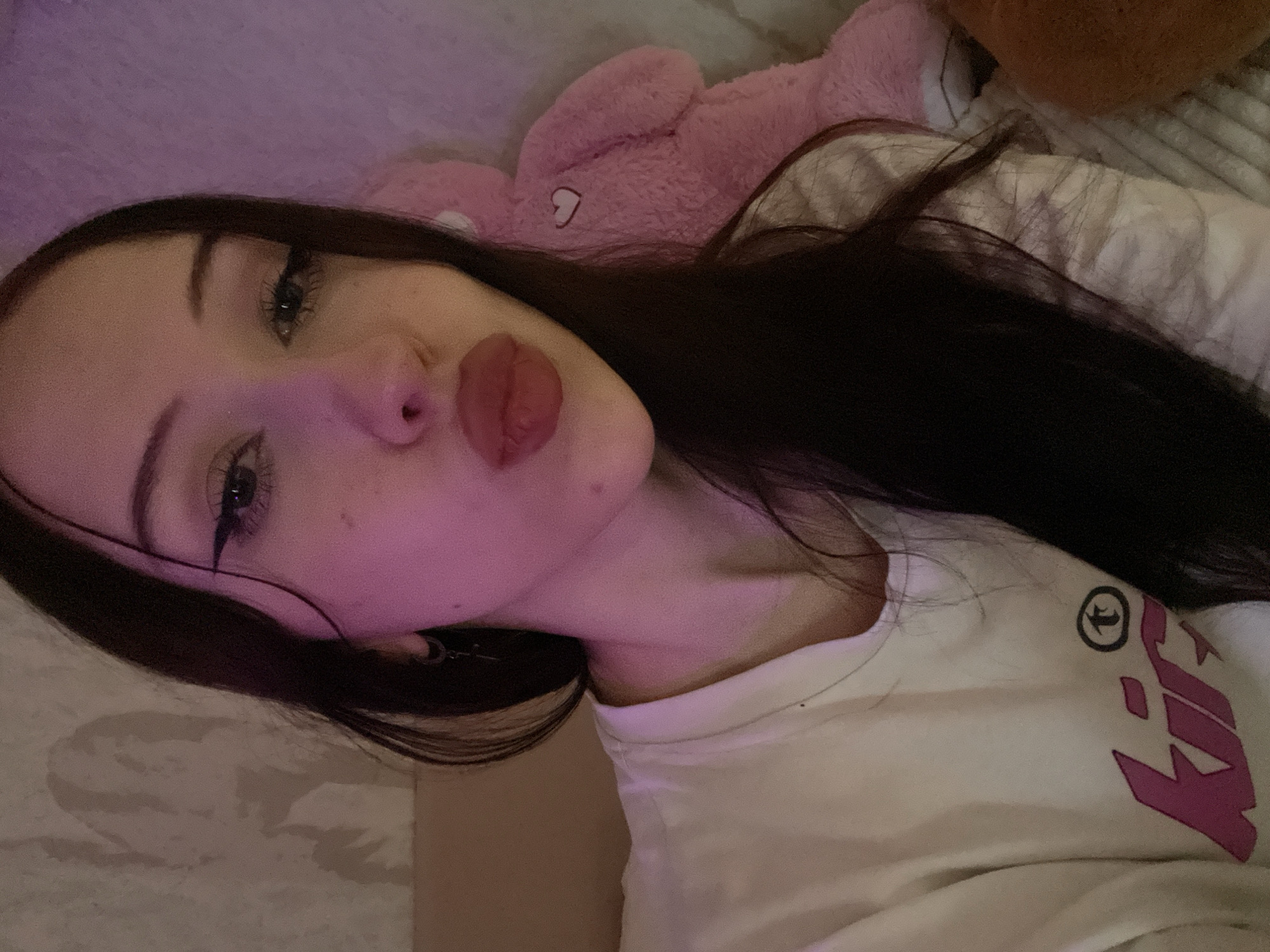 A young woman with long, dark hair and heavy eyeliner is taking a selfie. She is wearing a white t-shirt with a pink graphic design. She is puckering her lips. Behind her, a pink stuffed teddy bear with a heart on its chest is visible. The lighting in the room is a soft pink. The background appears to be a wall with a patterned wallpaper and a bed with a textured blanket. This image was taken in Votkinsk, Russia.