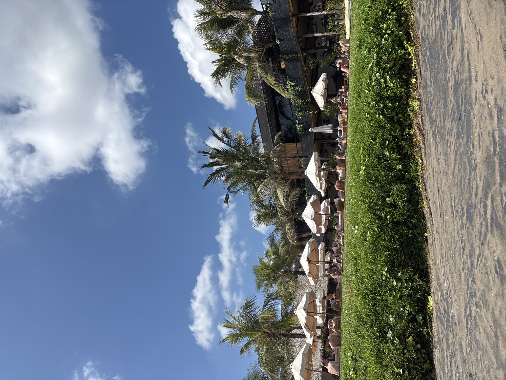 The image depicts a beach scene in North Kuta, Indonesia. The sky is blue with scattered white clouds. Palm trees line the beach, interspersed with white umbrellas shading rows of beach chairs occupied by people. Next to the beach is a restaurant. Adjacent to the beach chairs, a strip of green vegetation separates the beach from a sandy walkway. People are relaxing in the beach chairs.