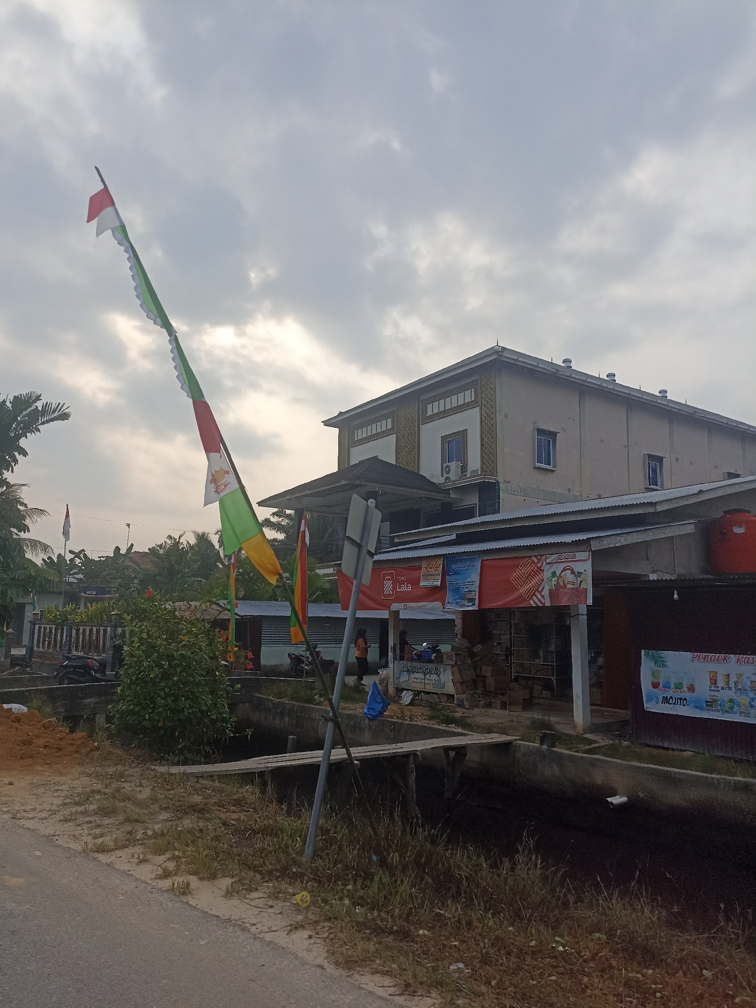 In Benteng Hulu, Indonesia, the scene depicts a cloudy day with a building complex as the dominant structure. A tall, thin building with multiple stories and a beige exterior featuring decorative gold patterns is partially visible. In front of it, a lower, single-story building with a corrugated metal roof houses several small shops or stalls, indicated by signage. One sign, in red and white, displays text that appears to be promotional. Orange water tanks are visible on top of this lower building. A paved road runs in the foreground, with a dry, grassy area and a small, narrow canal adjacent to it. A wooden plank serves as a makeshift bridge across the canal. A decorative flag, predominantly green and white with red accents, is prominently displayed on a pole to the left of the frame, extending towards the sky. Several motorcycles are parked near the shops. The environment suggests a mixed-use area, possibly a commercial district with residential elements nearby.