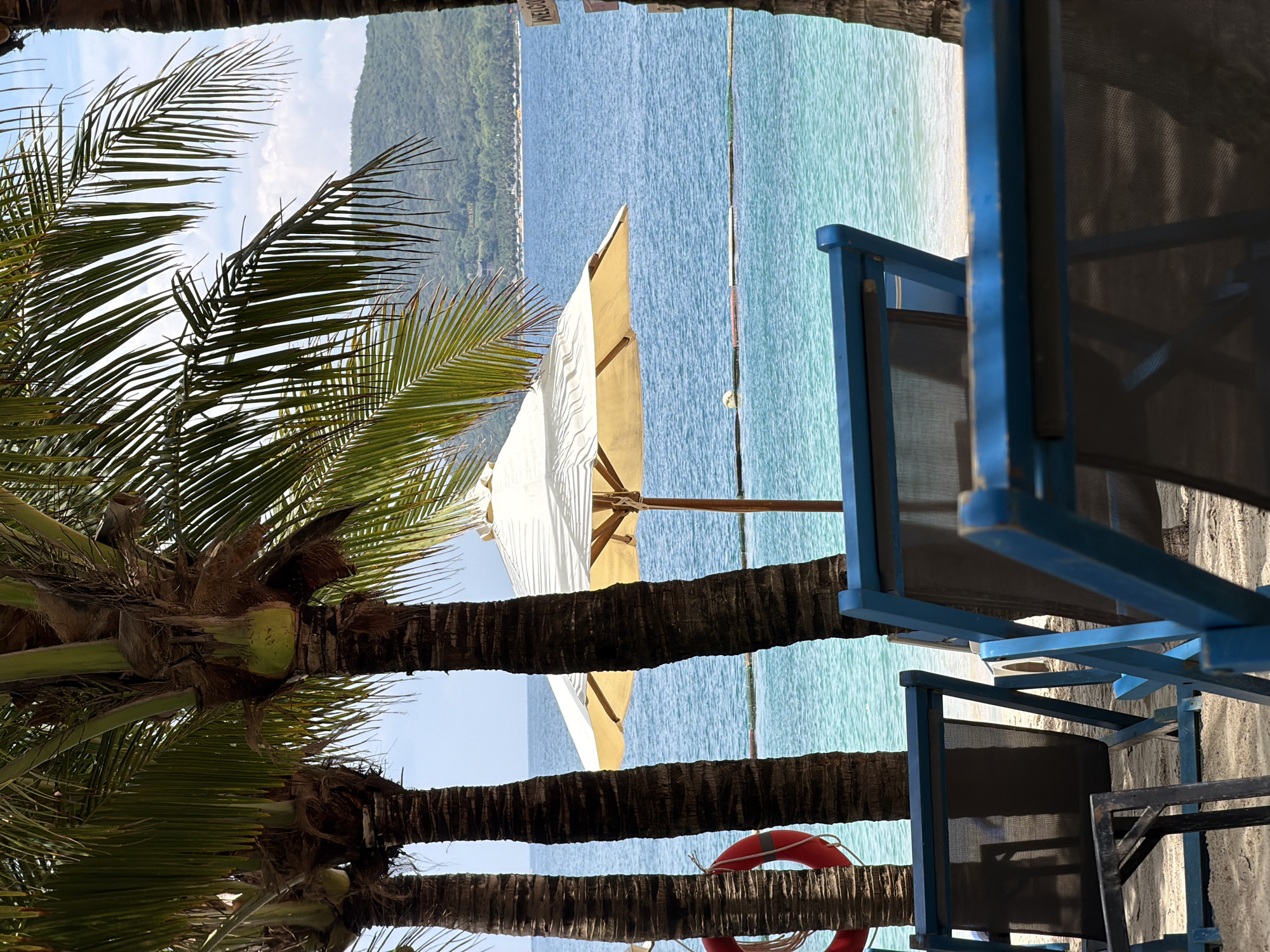 A beachfront scene in Nam Nha Trang Ward, Vietnam is depicted. The image is framed by the trunks of palm trees and the structure of blue beach chairs. Beyond the palm trees and beach chairs, a white beach umbrella stands on a sandy beach beside the ocean. The ocean is a gradient of light blue and darker blue, and land is visible in the distance. A red life preserver rests against a palm tree trunk.
