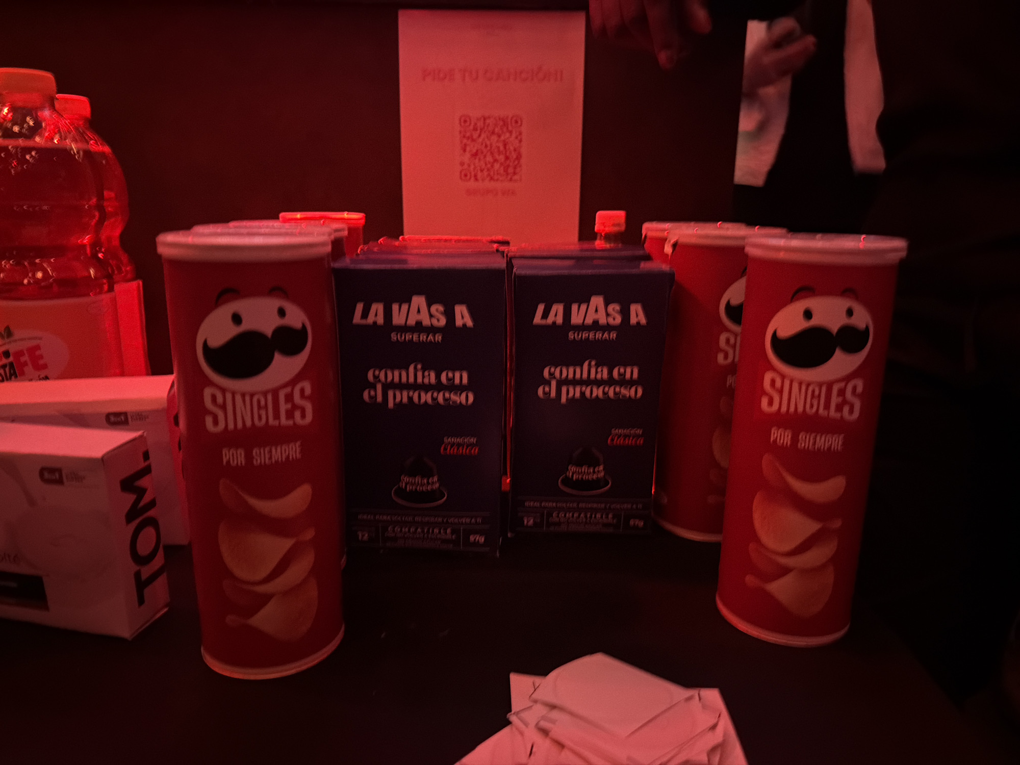 A dimly lit scene under red lighting showcases a table with various items. On the left are stacked bottles of a beverage labeled "STA FE" and a box with "TOM." printed on it. Two cans of "Pringles Singles" stand prominently next to two dark blue boxes labeled "LA VAS A SUPERAR," which translates to "You'll Overcome It," alongside the text "confia en el proceso," meaning "trust the process." A white sign in the background reads "PIDE TU CANCION," meaning "Request Your Song," with a QR code beneath. A person is visible in the background, partially obscured. In the foreground are white napkins, and the table appears to be the main focal point of the image. Location is not evident from the content of this image.