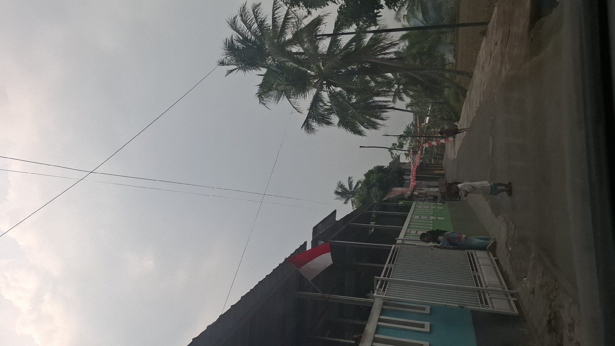 In Sukasari, Indonesia, a street scene unfolds under a cloudy sky, with power lines crisscrossing overhead. To the left, a building with a dark, slanted roof is bordered by a light-colored metal fence. An Indonesian flag, red and white, flies from the building's eaves. On the street in front of the building, a woman and a child stand near the fence. Further down the street, a child in a light-colored shirt and blue shorts stands with their hands in front of them. Beyond this child, a darker-skinned figure runs away from the viewer. The street curves to the right, flanked by palm trees and other greenery. A field is visible in the distance, and small red and white flags or banners are strung along the buildings on the right side of the street, suggesting a local celebration or observance. The overall atmosphere is overcast and somewhat hazy.