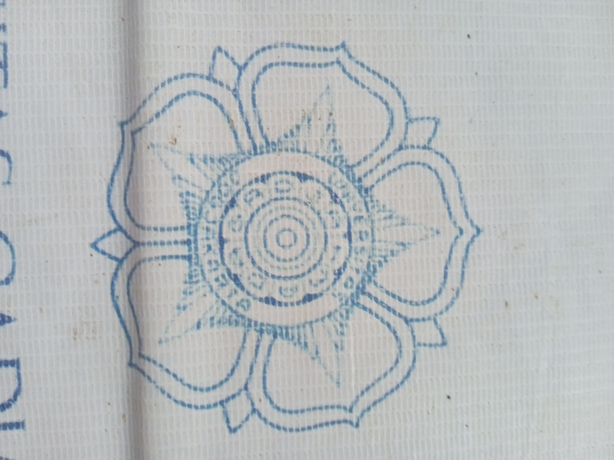 A blue, circular, stylized floral or mandala pattern is imprinted on a white, woven textile. The pattern features an outer perimeter of eight connected, rounded petal shapes, an inner eight-pointed star motif, and multiple concentric circles with dashed and solid line details culminating in a small central circle. The blue coloration, exhibiting minor variations in density, appears to be composed of short dashed segments, consistent with a stamp or print. The underlying fabric is off-white, displaying a prominent, uniform grid-like weave texture. Sparse dark particulate matter is scattered across the textile surface. This image depicts an object located in Teupinjok, Indonesia.