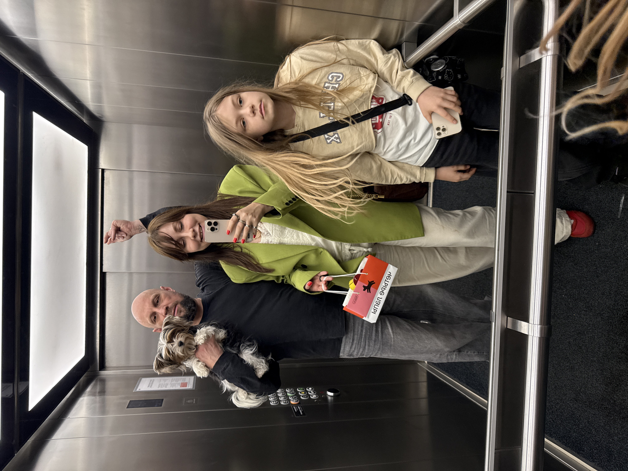 The image shows three people and a dog in an elevator. A bald man with a beard is holding a small black and white dog. A woman is holding a smartphone and taking a selfie. A young girl is standing next to her. The woman is holding a red and white paper bag with lettering on it. The elevator has stainless steel walls and a black floor. There is a window with light coming through it. Based on the writing on the bag, the location is Moscow, Russia.