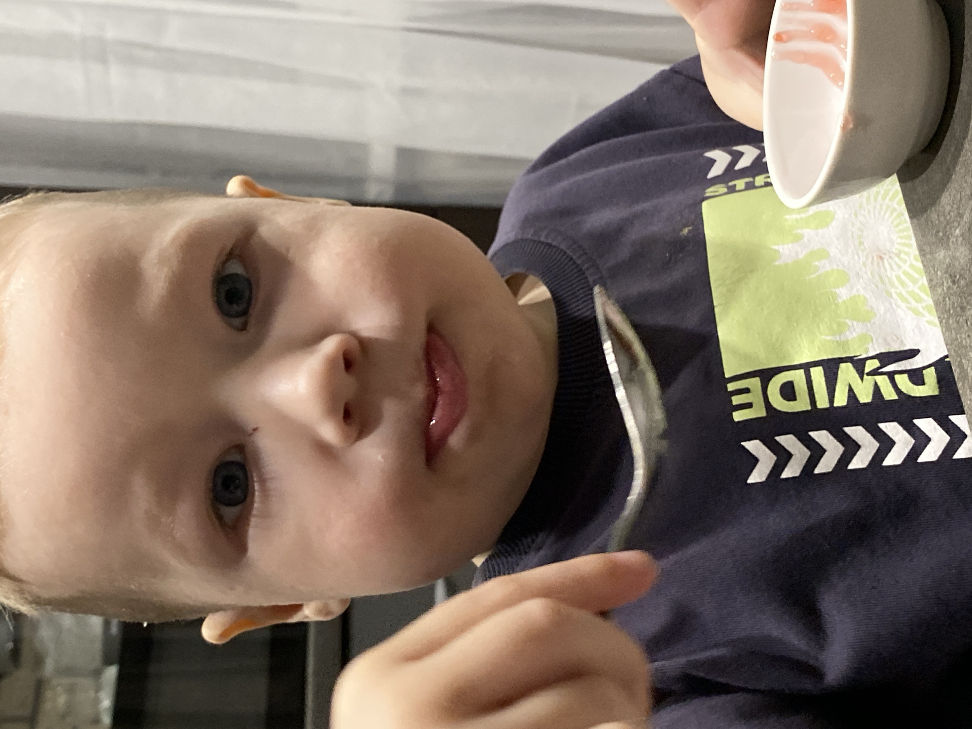 A close-up, slightly rotated image features a young child, likely a boy, with fair skin, light-colored hair, and bright blue eyes, engaged in eating. The scene takes place indoors, with the location context being Sochi, Russia. The child holds a silver spoon, and a small white bowl, containing remnants of a reddish-pink substance like fruit puree or yogurt, is visible near their hand. The child's expression is focused, with a soft, slightly pouting look.
They are dressed in a dark blue long-sleeved shirt that features a prominent lime green and white graphic design. Visible text on the shirt includes "WIDE" (appearing inverted due to the image's rotation) and a partial "STR," accompanied by multiple white chevron arrow patterns. The background is softly blurred, showing horizontal lines that suggest an indoor setting, possibly a home dining area or kitchen, under ambient indoor lighting.