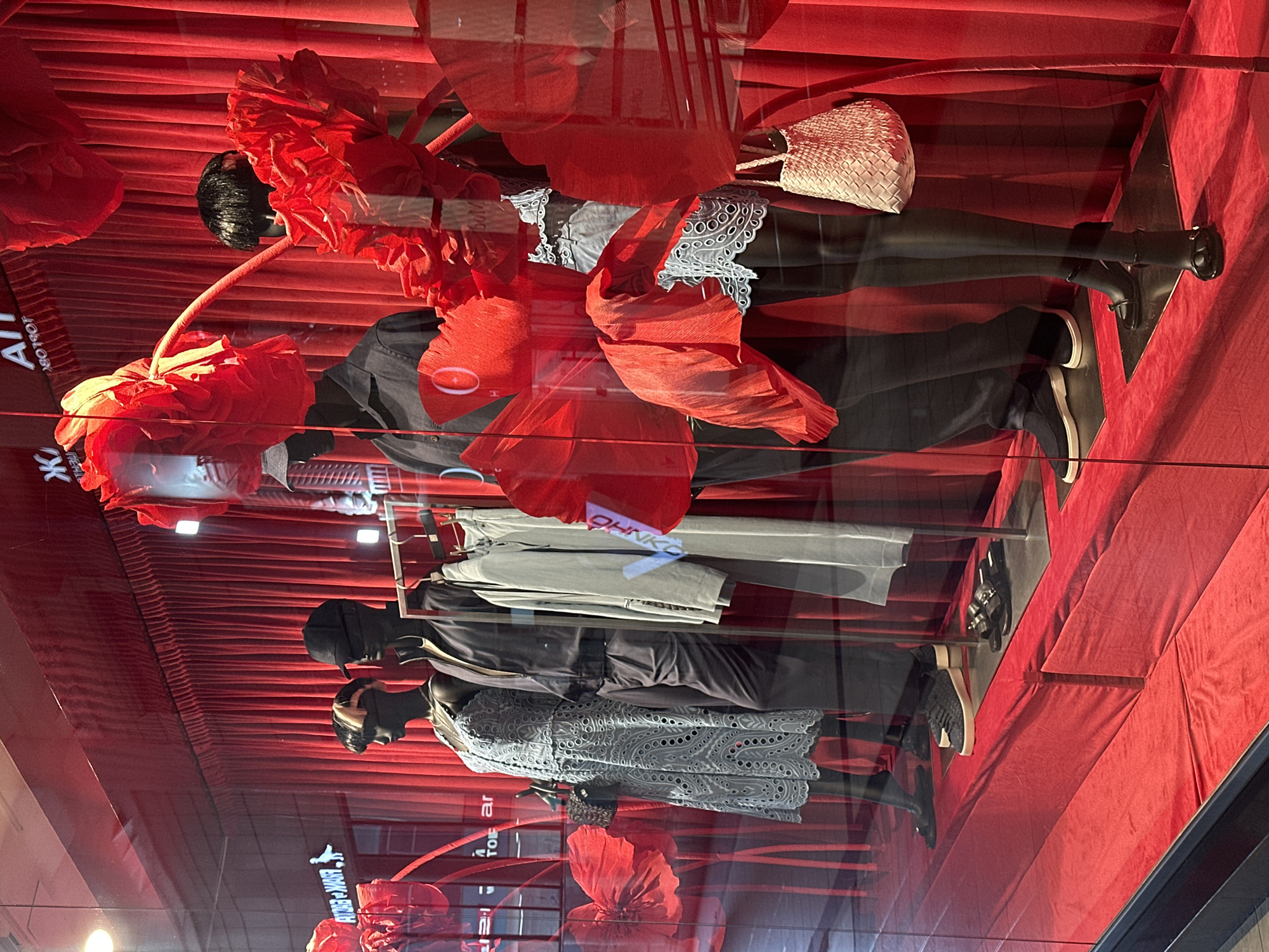 A display window on the ground floor of a retail establishment in Sochi, Russia, features mannequins dressed in fashionable clothing. The display is dominated by a vibrant red color scheme, with red velvet curtains serving as the backdrop. Large, abstract red floral decorations are strategically placed throughout the window, adding a dramatic and artistic flair.
In the foreground, several mannequins are positioned. The leftmost mannequin, a female, wears a light blue, intricate lace dress and carries a black handbag. Next to her, a male mannequin is dressed in a dark grey button-down shirt and dark trousers. To his right, another male mannequin wears a dark grey zippered jacket over a white shirt and dark trousers. Further to the right, a female mannequin sports a white lace top and dark bottoms. Another male mannequin is visible on the far right, wearing dark trousers.
On the floor of the display, various footwear is arranged. Black slip-on sandals are visible on the left, followed by black woven sneakers with white soles. Further to the right, black sandals with buckle details are displayed. A woven light-colored handbag hangs from the rightmost female mannequin's arm.
The mannequins are arranged to showcase a coordinated collection of apparel, with a focus on stylish and modern aesthetics. The overall impression is one of sophisticated retail display, utilizing bold colors and artistic elements to attract attention. Reflections of the environment outside the window, including street lights and possibly other buildings, are visible on the glass.