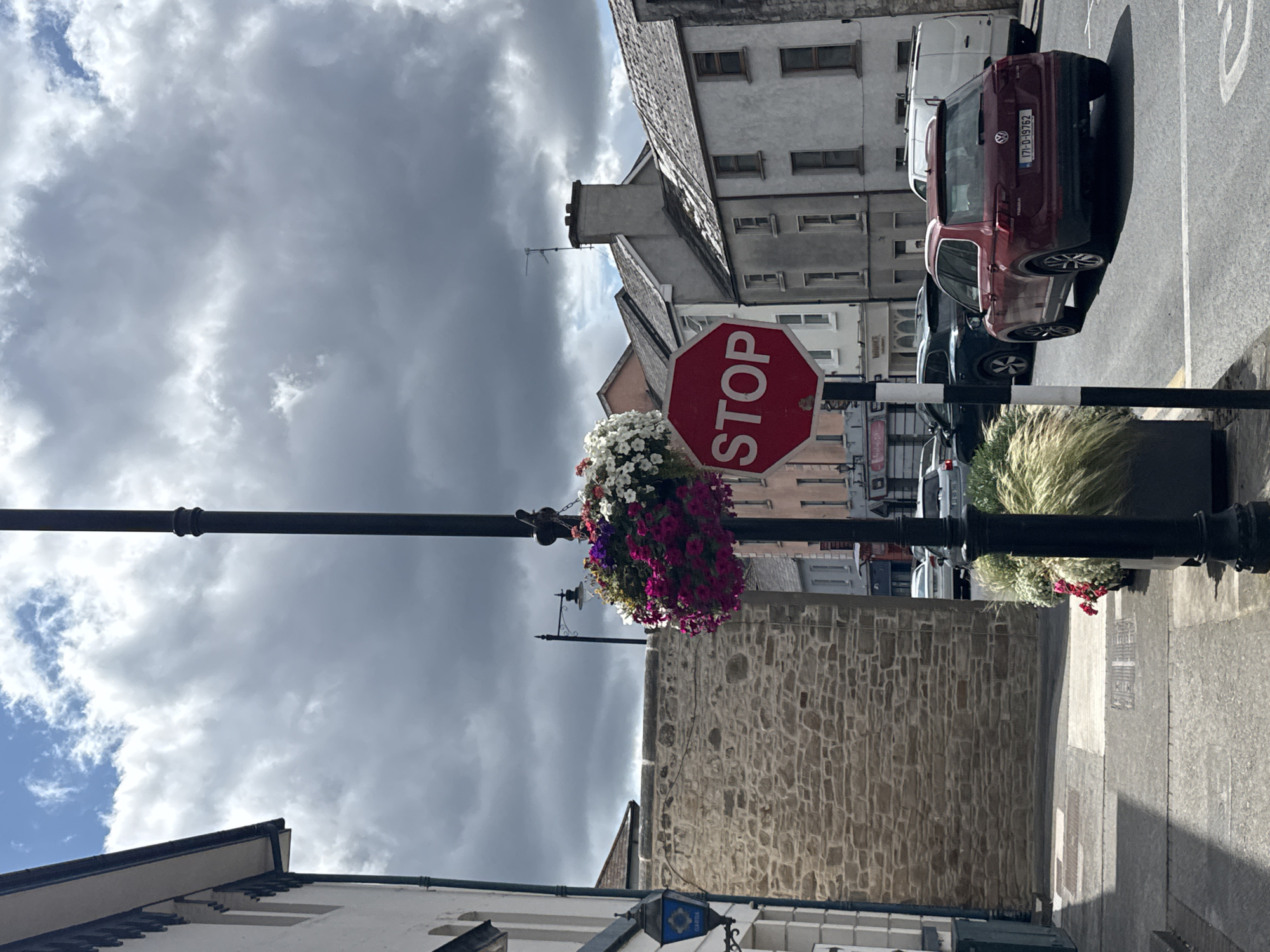 The image depicts a street scene in Clonmel, Ireland, with a focus on a black lamppost adorned with hanging baskets filled with colorful flowers, predominantly pink and white petunias. A prominent red octagonal stop sign is attached to the lamppost, facing the viewer. In the foreground, a portion of a stone wall is visible to the left, with a blue Garda (Irish police) lamp mounted on a building's facade. The street is paved, and a dark red Volkswagen Tiguan is driving away from the viewer, displaying an Irish license plate that reads "171-D-19762." Other vehicles, including a white van and a dark-colored SUV, are also present on the road. The background features a row of buildings with pitched roofs, characteristic of Irish architecture, under a dramatic sky filled with heavy, gray clouds and patches of blue. The overall atmosphere suggests an overcast day in an urban setting.